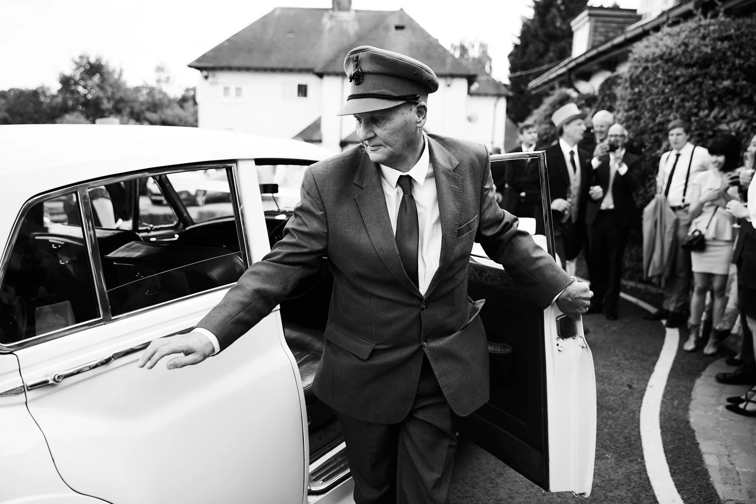 A man in a vintage military-style hat and a suit gets out of a classic white car surrounded by people dressed in formal attire, at what appears to be a formal outdoor event.