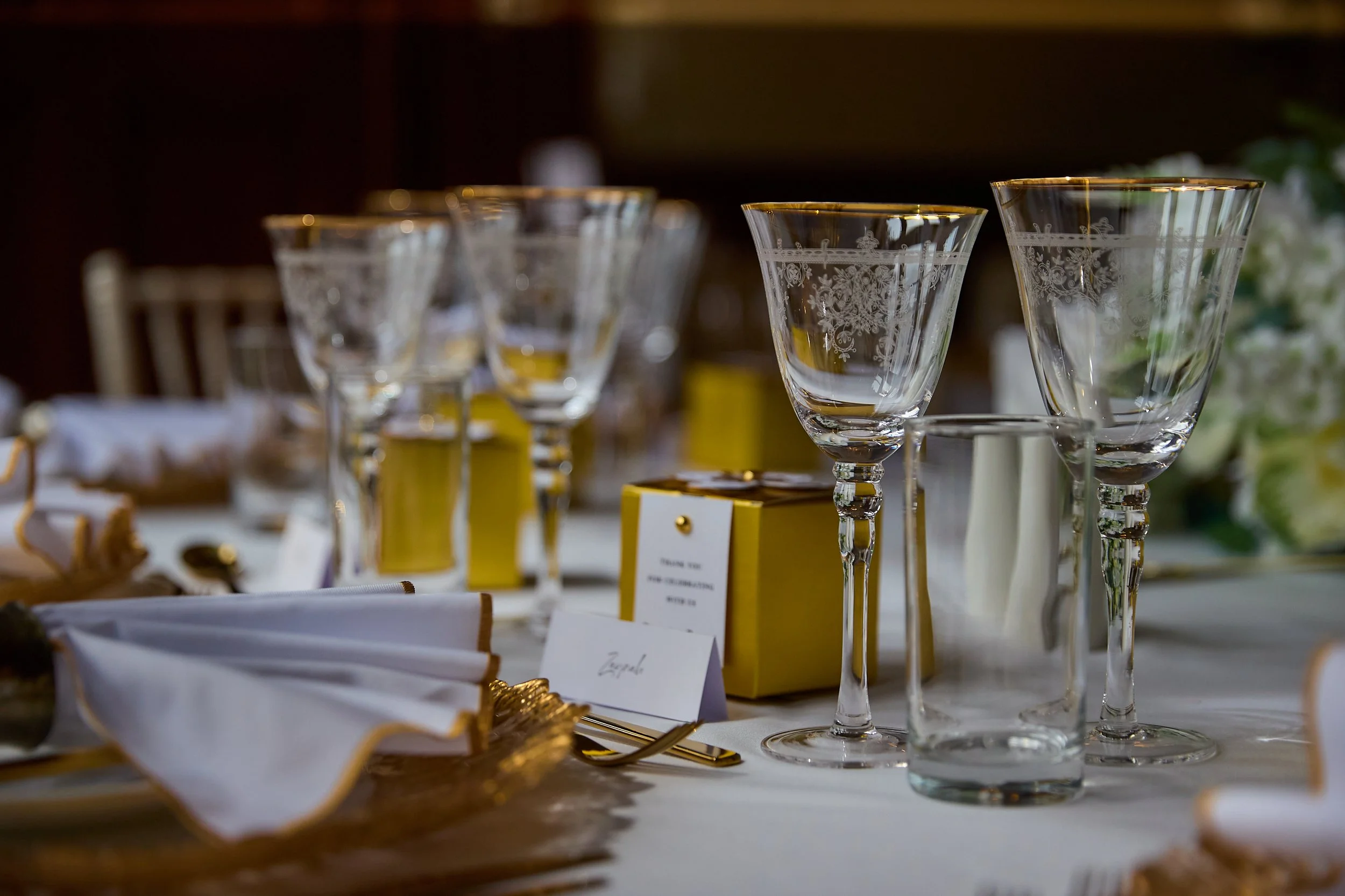 Elegant table setting with engraved glassware, white napkins with gold trim, and yellow gift boxes on a white tablecloth for a formal event or celebration.