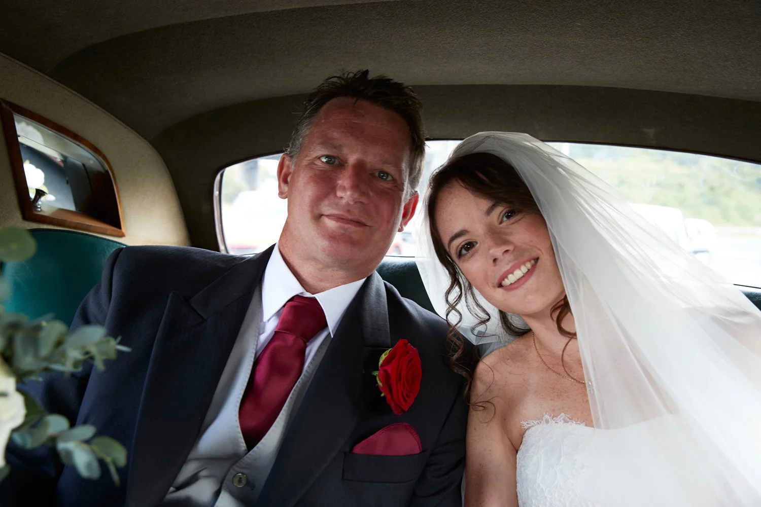 A newlywed couple sitting in the back of a vehicle, smiling at the camera. The groom wears a suit with a red tie and a red rose boutonniere. The bride is dressed in a white wedding gown with a veil.