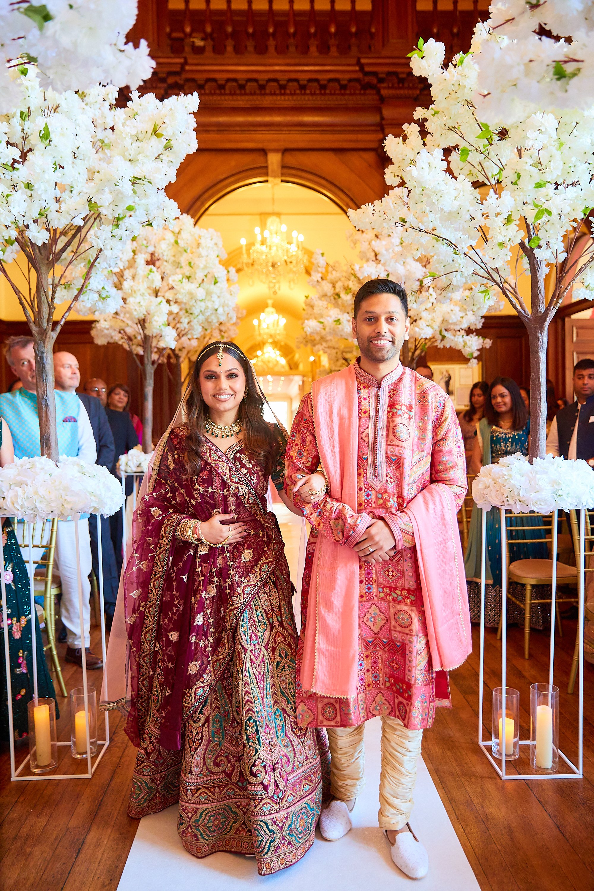 A couple in traditional Indian attire standing together in a decorated wedding venue with white flowers and candles, surrounded by guests.