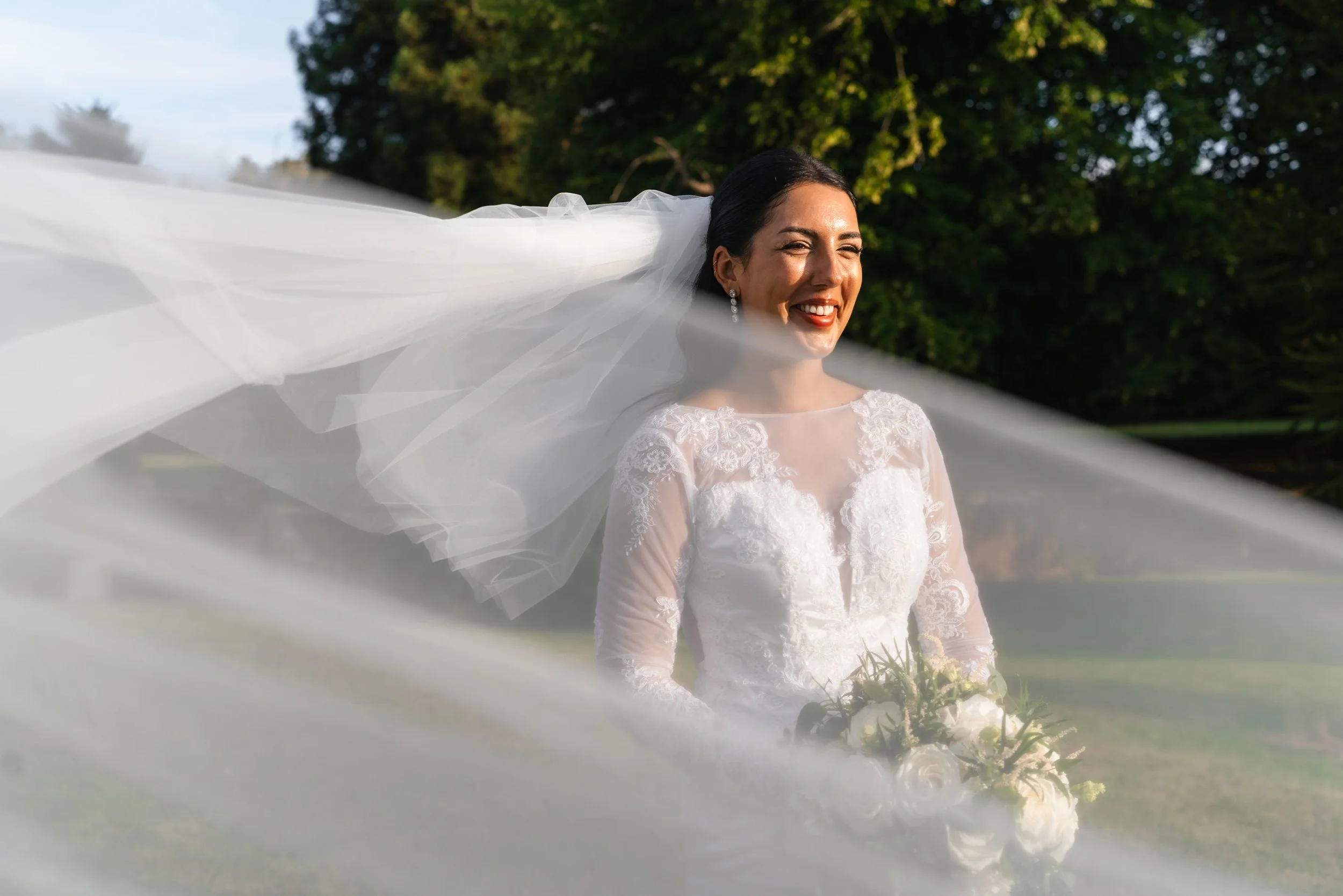 Bride in a white lace wedding dress holding a bouquet, smiling outdoors with a flowing veil and lush green trees in the background.
