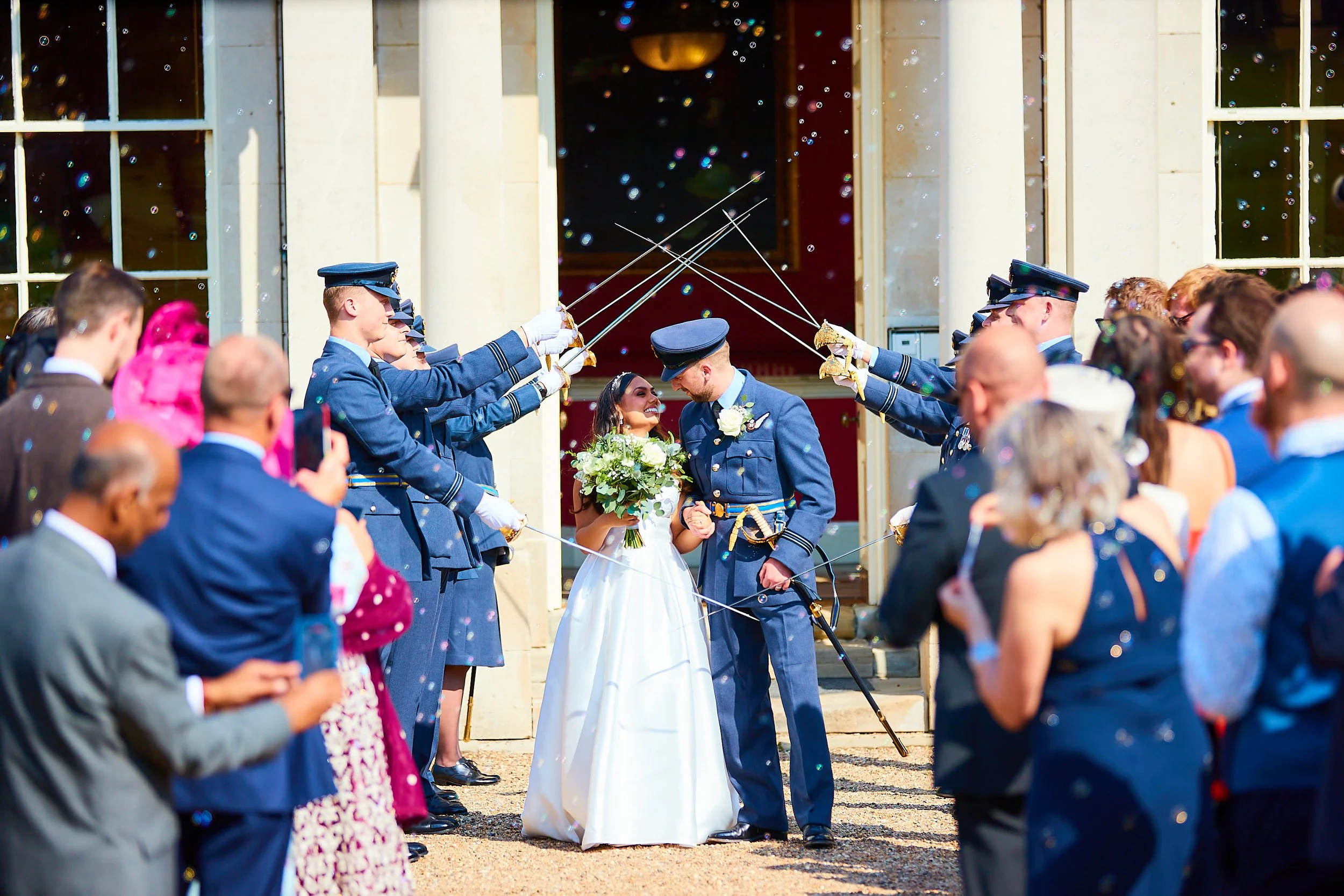 A bride and groom holding hands, standing amidst uniformed guards who are saluting them with swords forming an arch, in front of a building with columns, during a wedding celebration.