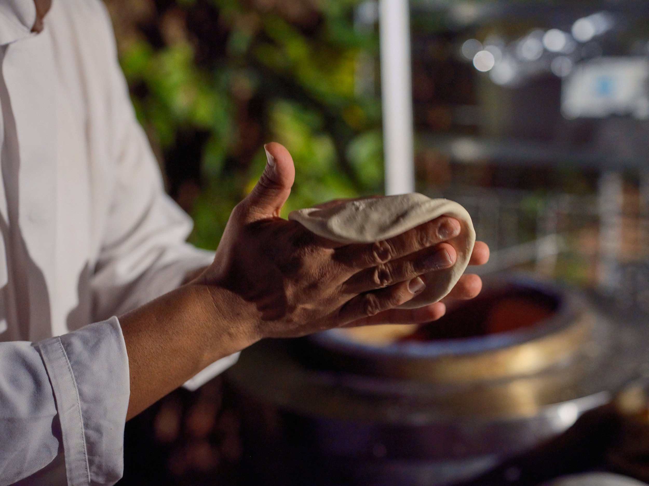 Person preparing traditional flatbread dough outdoors at sunset, with a stack of flatbreads on a tray in the background.
