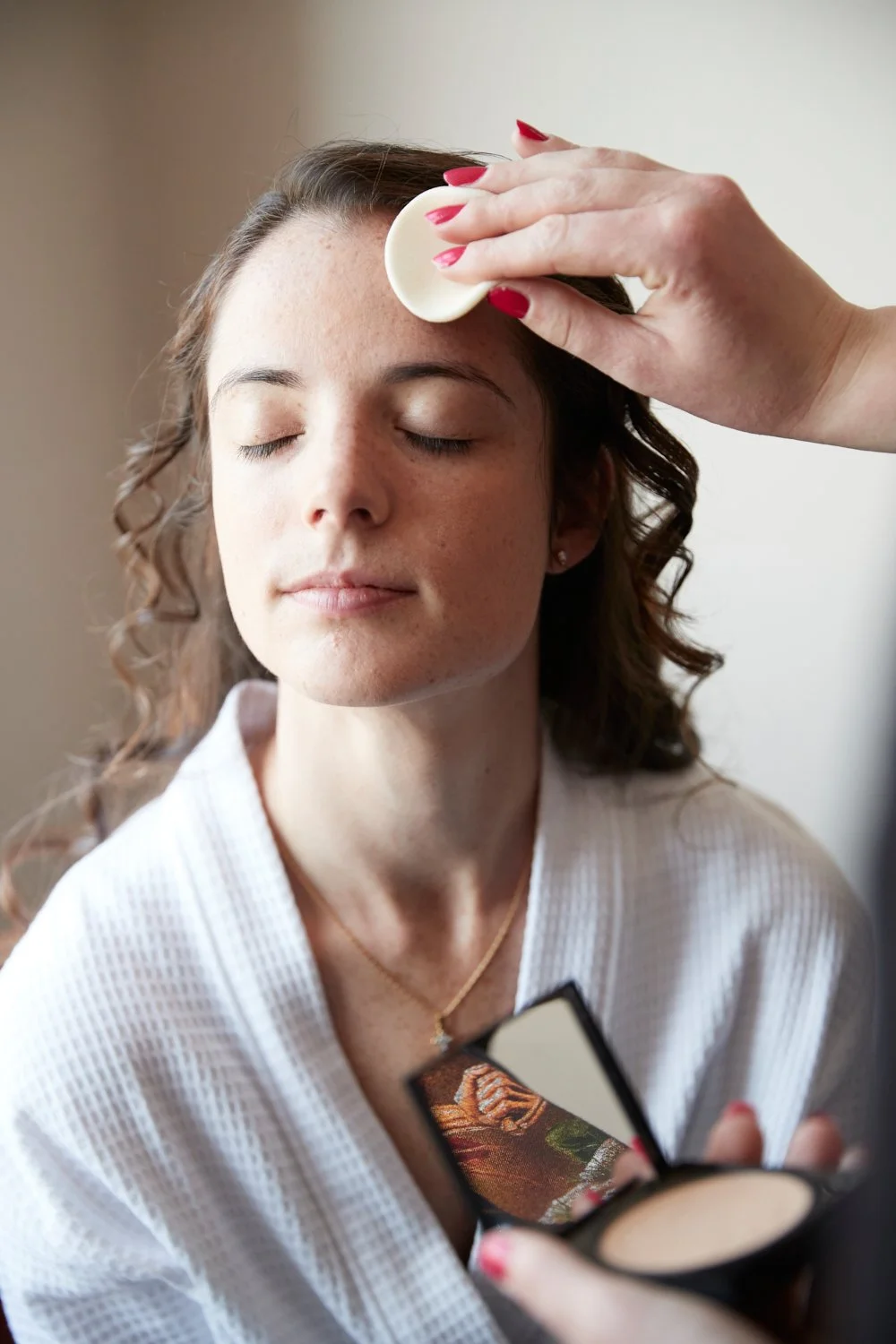 Woman with closed eyes having her forehead, drawn as a part of a beauty or skincare routine, gently wiped with a round pad by another hand. The woman wears a white textured robe and a gold necklace, with a makeup compact open on her lap.