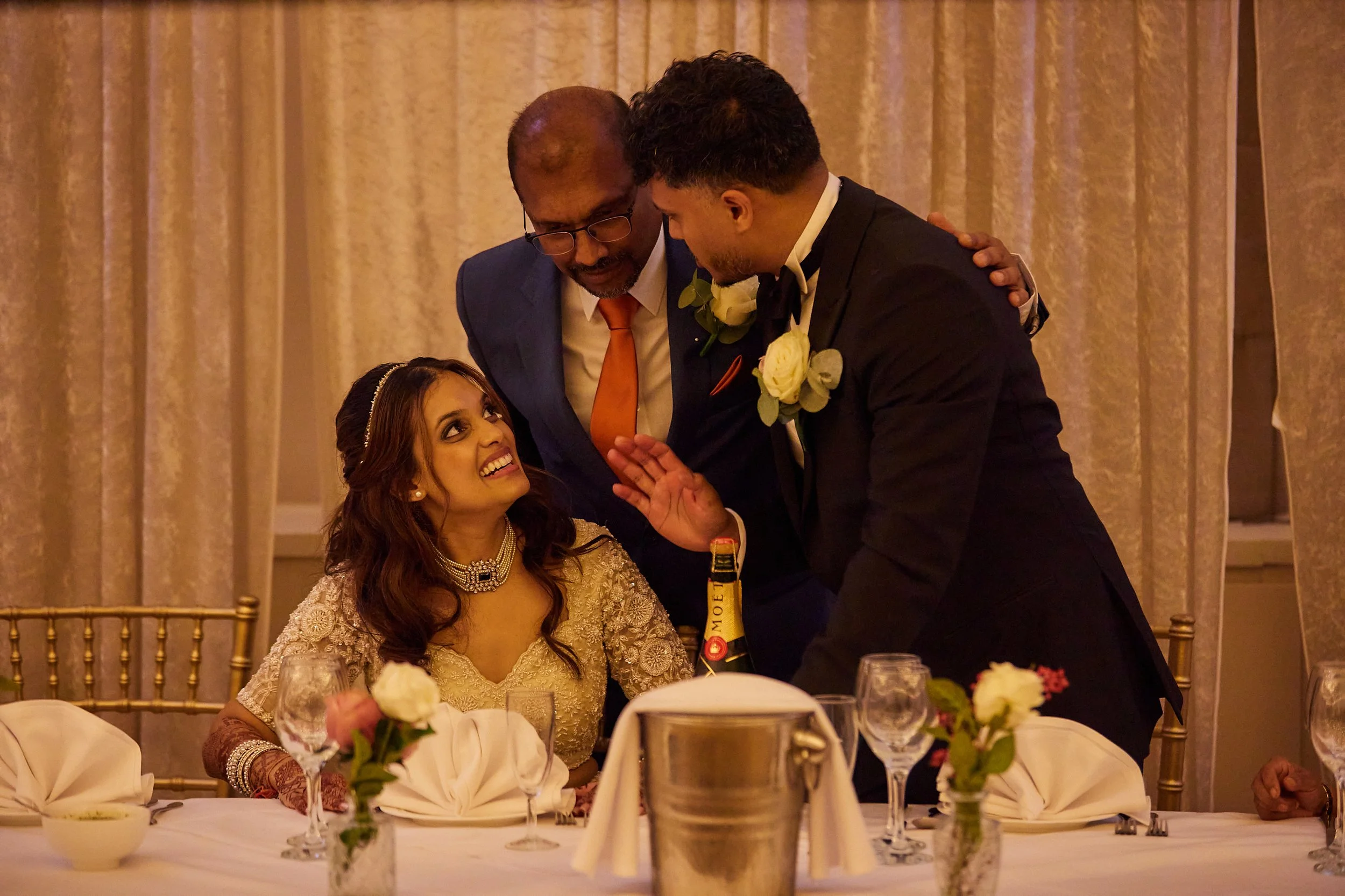 A woman in a gold dress and jewelry sitting at a wedding reception table, smiling at a man in a tuxedo and boutonniere, who is speaking to her. An older man in a suit and tie stands behind them, leaning in. The table has flowers, glasses, and a champ