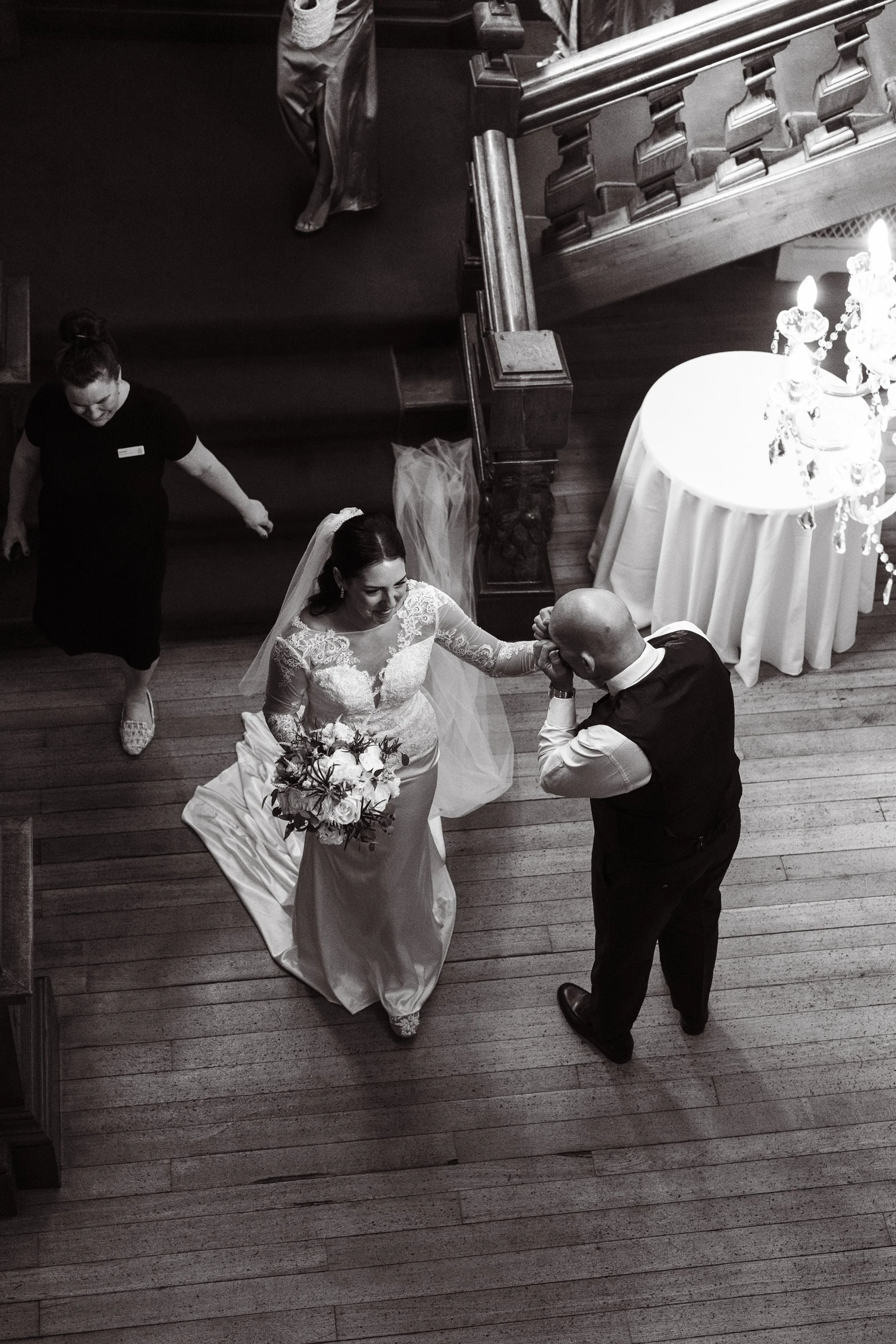 Black and white photo of a bride with a bouquet of flowers sharing a kiss with a bald man in formal attire, likely at a wedding venue, with a woman walking in the background. The photo is taken from above, showing a wooden staircase and a table with 