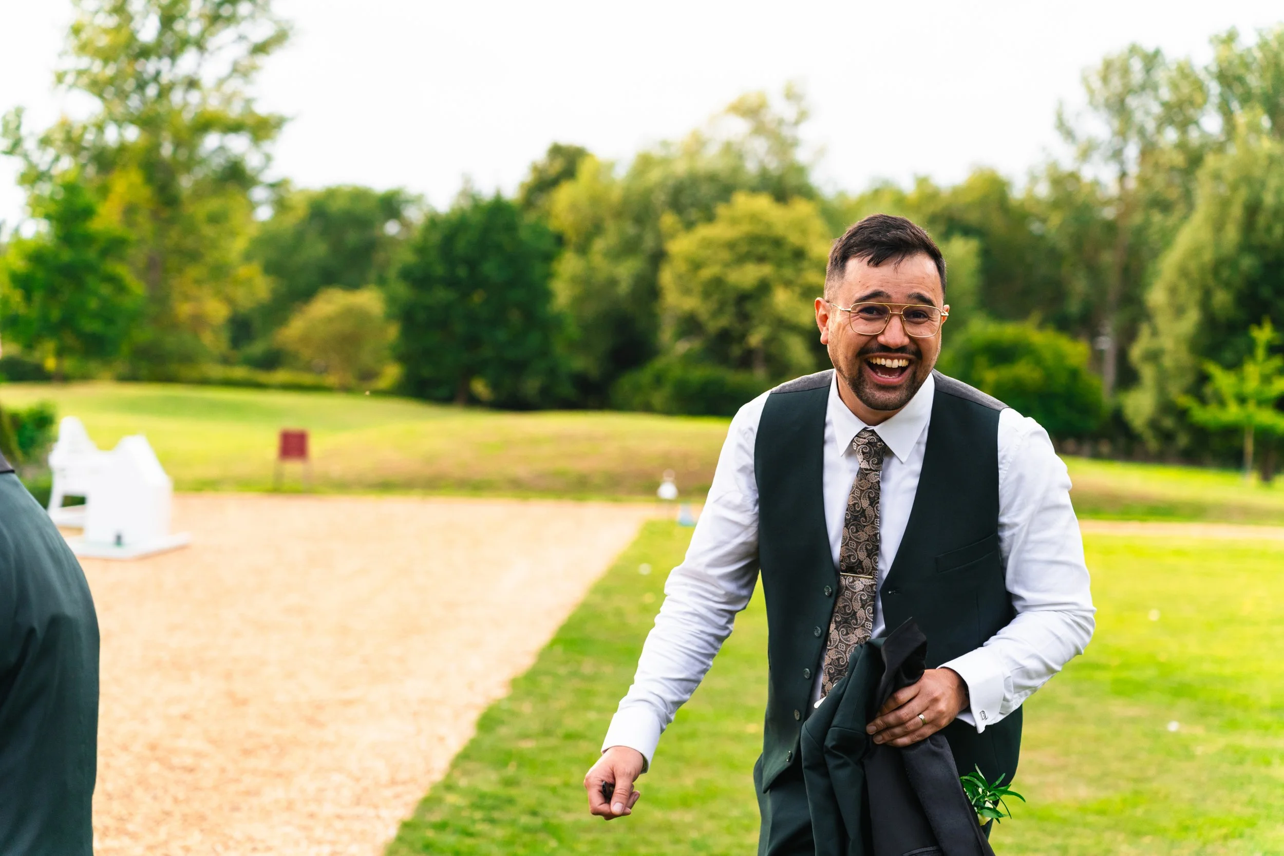 A man dressed in formal attire, smiling and laughing outdoors, holding a jacket, with a lush green park in the background.