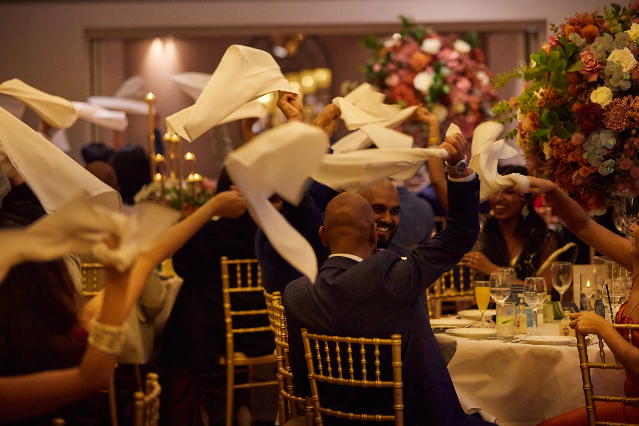 Guests at a wedding celebration waving white napkins with joyful expressions, surrounded by floral decorations and elegant table settings.