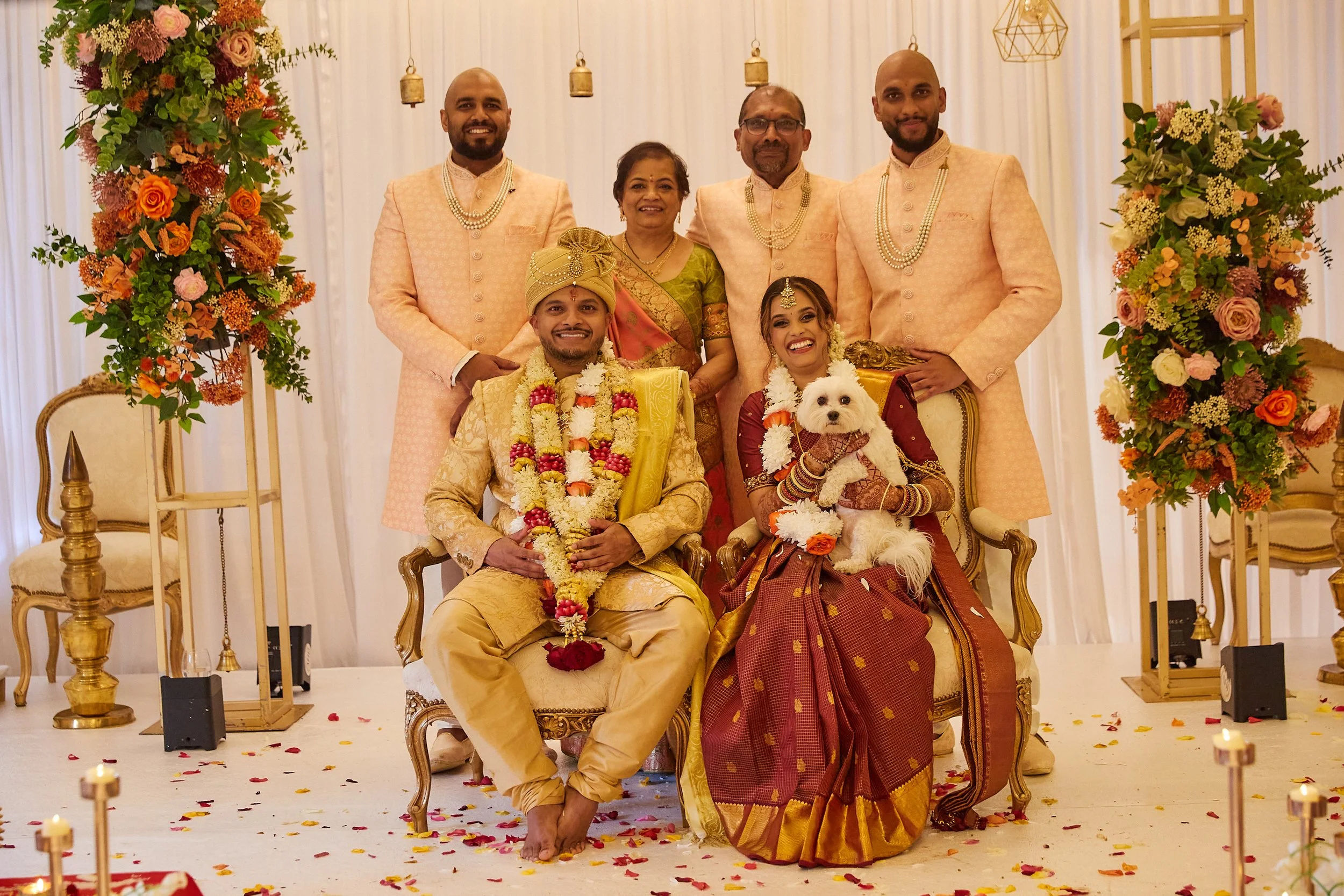 A traditional Indian wedding celebration with the bride and groom and their family members dressed in colorful attire, sitting and standing in a decorated venue with flowers and ornaments.
