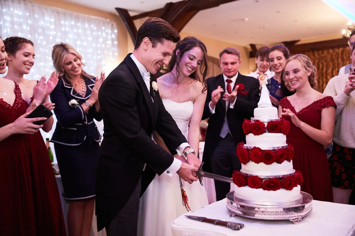 A newlywed couple cuts their wedding cake surrounded by smiling friends and family at a celebration.