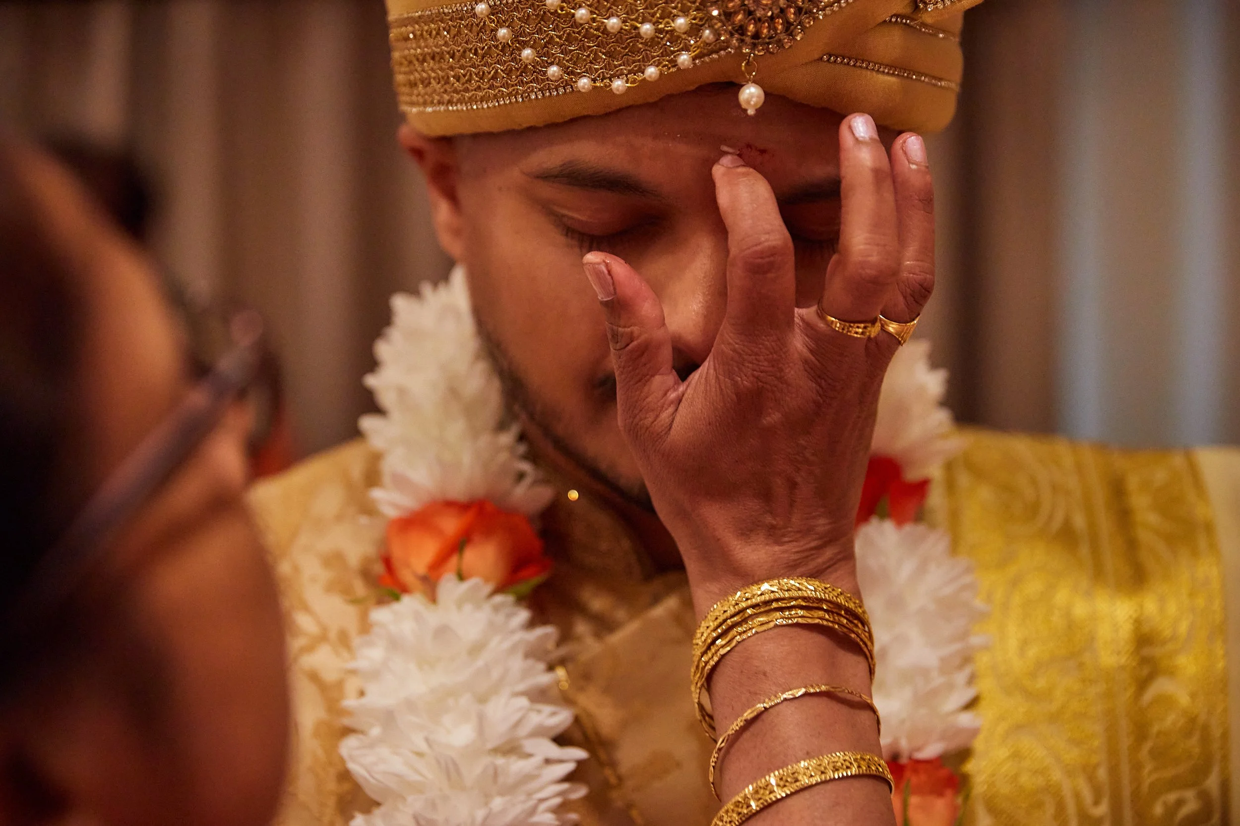 A man dressed in traditional attire with gold jewelry, holding his face in prayer or reflection, engaged in a cultural or religious ceremony.