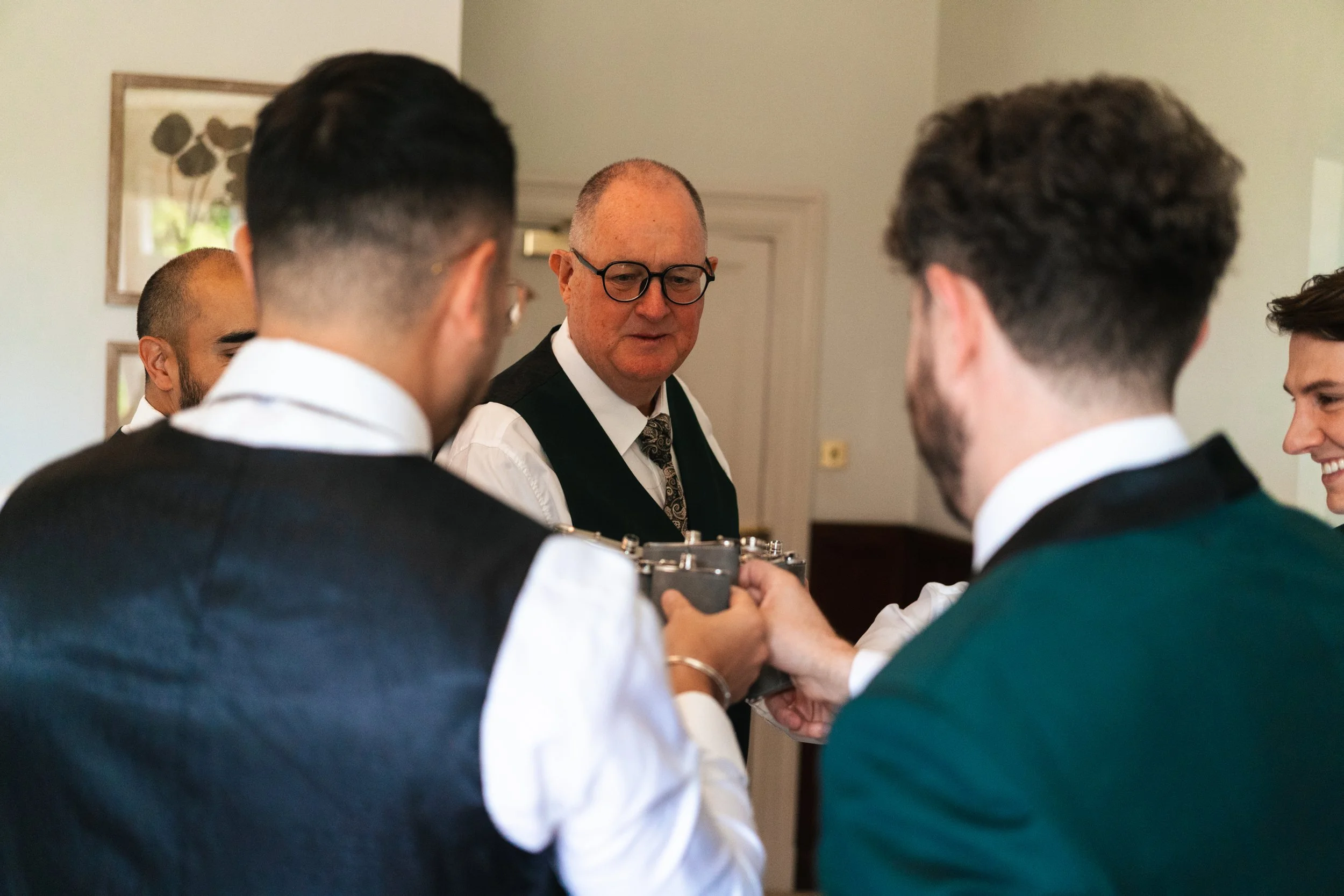 A group of men in formal attire, with one man in the middle holding a camera, gathered around and smiling in a well-lit room.
