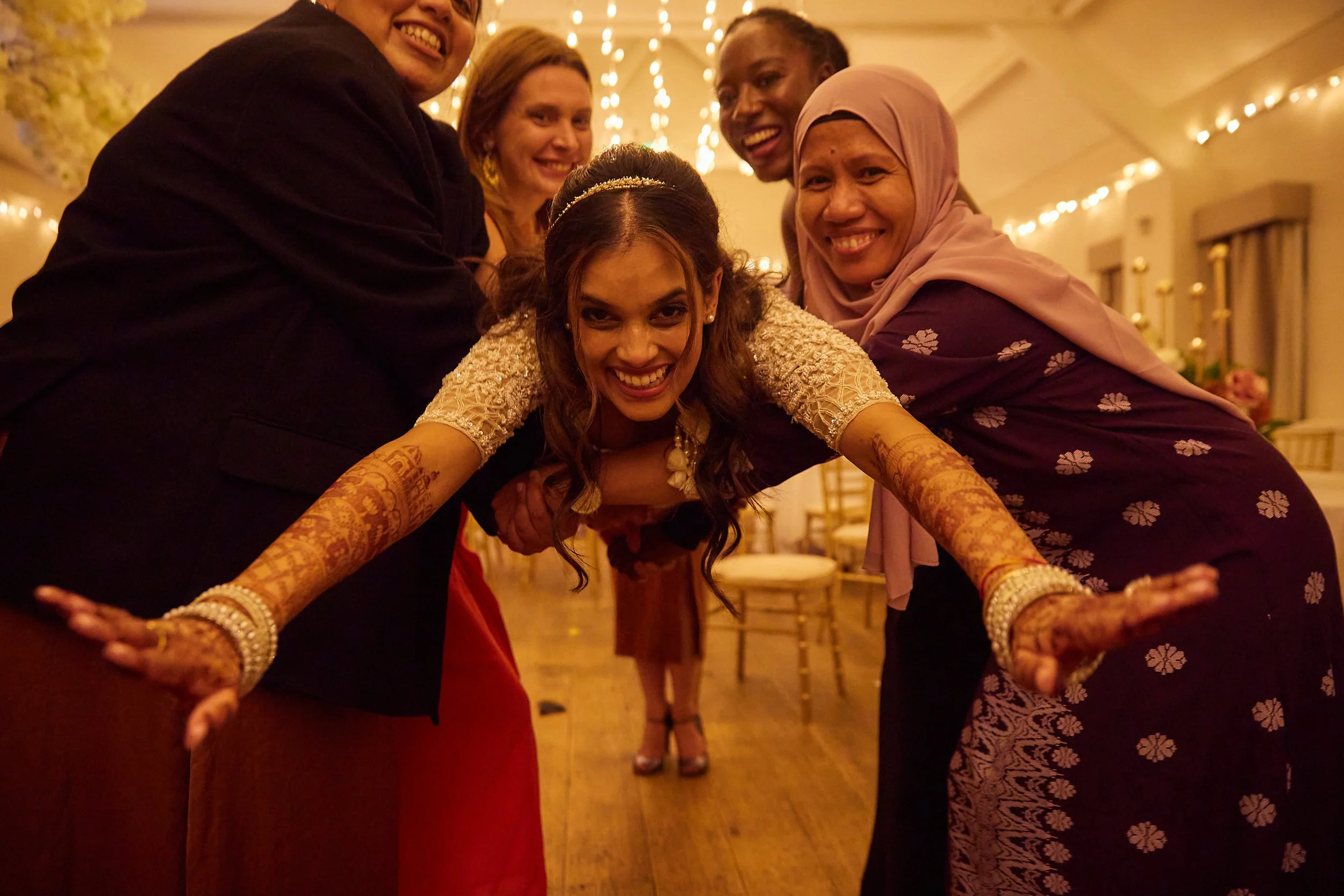 Group of six women, including a bride, smiling and posing at a celebration or wedding in a decorated venue with string lights and chairs in the background.