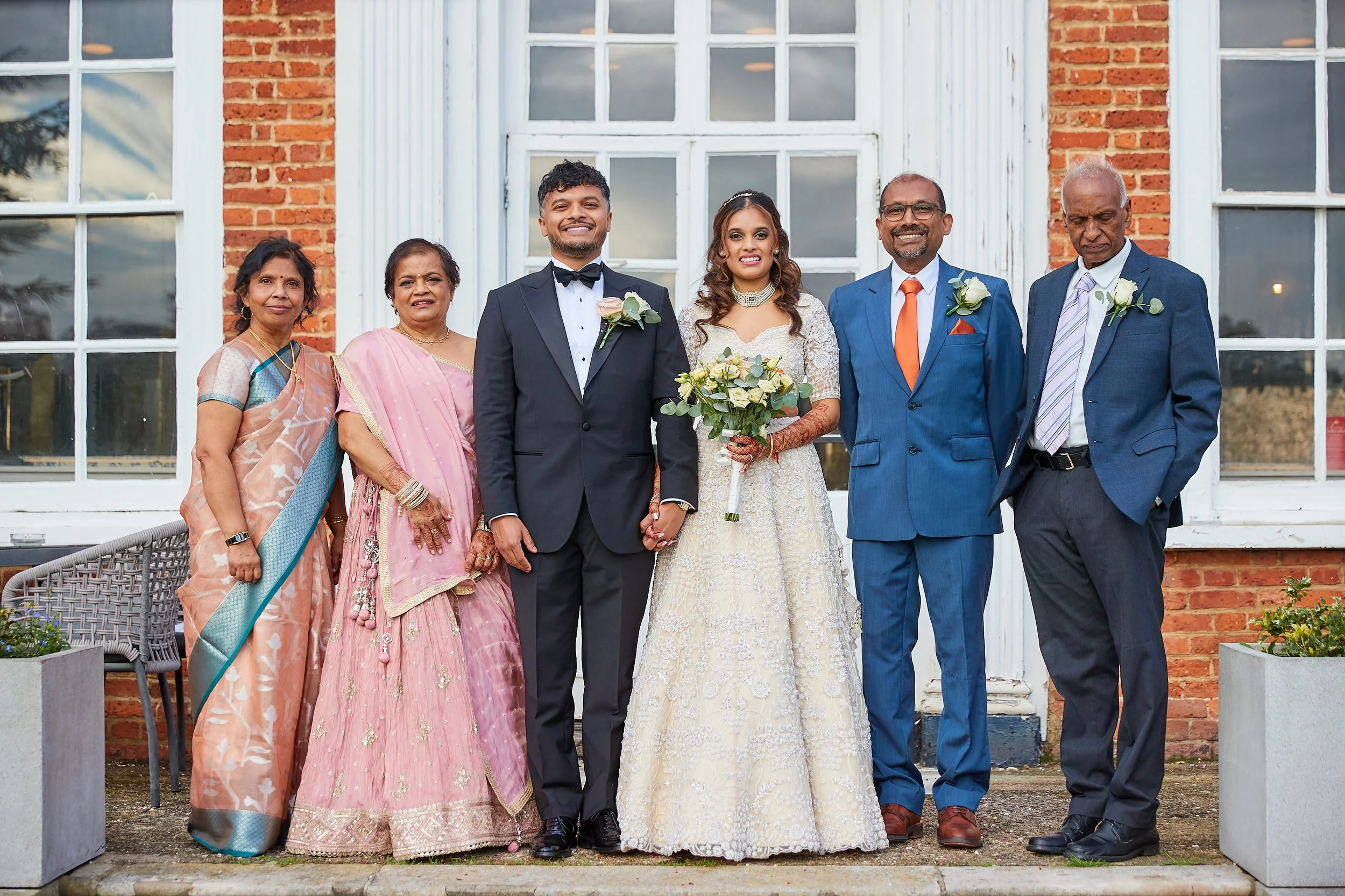 A wedding group photo featuring six people standing outside in front of a brick building with large windows. The bride in a white lace gown holding a bouquet, and the groom in a black tuxedo with a bow tie, are in the center. Flanking them are family