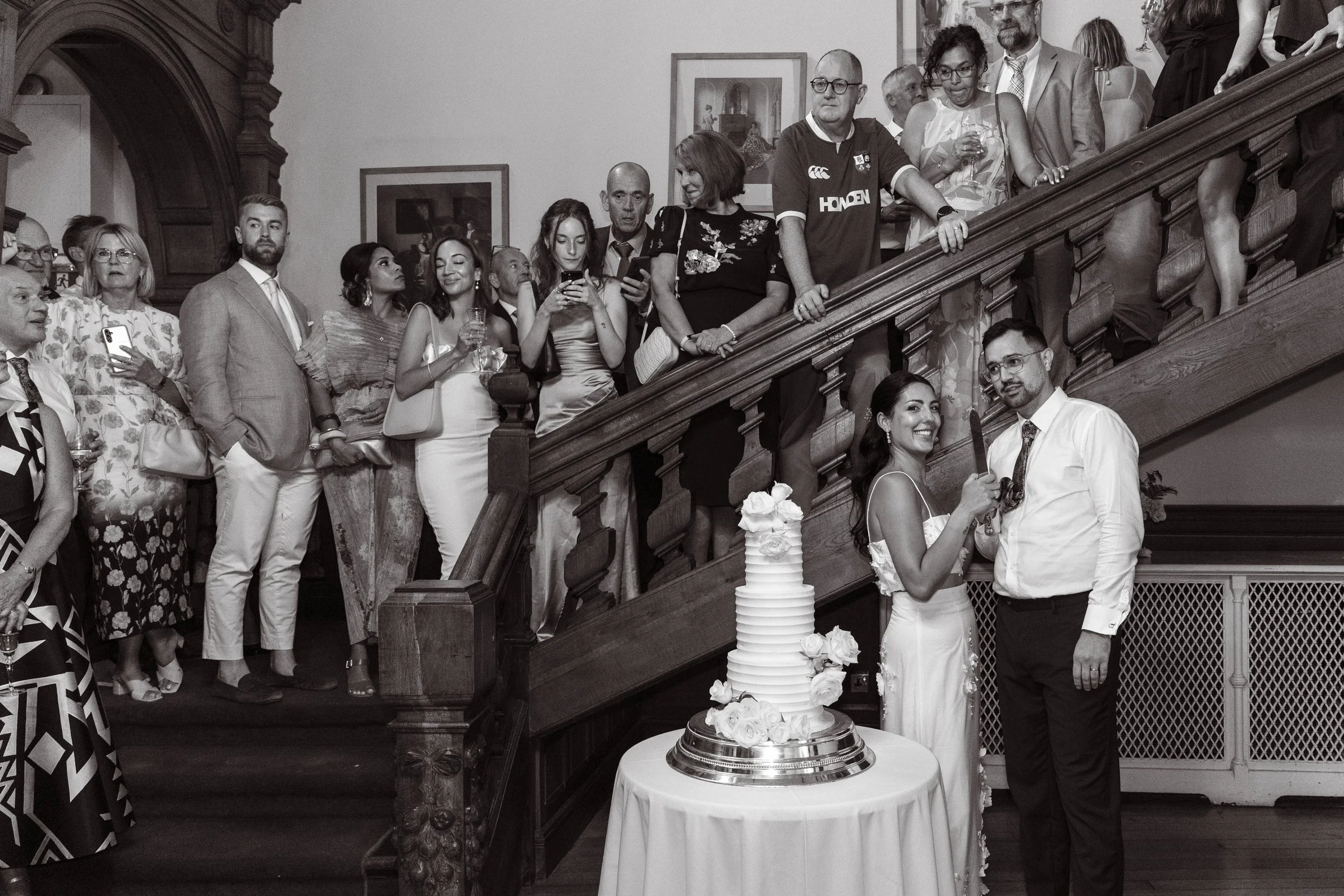 A wedding reception with a bride and groom standing in front of a tall wedding cake. The bride is smiling and holding a knife, ready to cut the cake. Guests are gathered on the staircase behind them, watching and taking photos.
