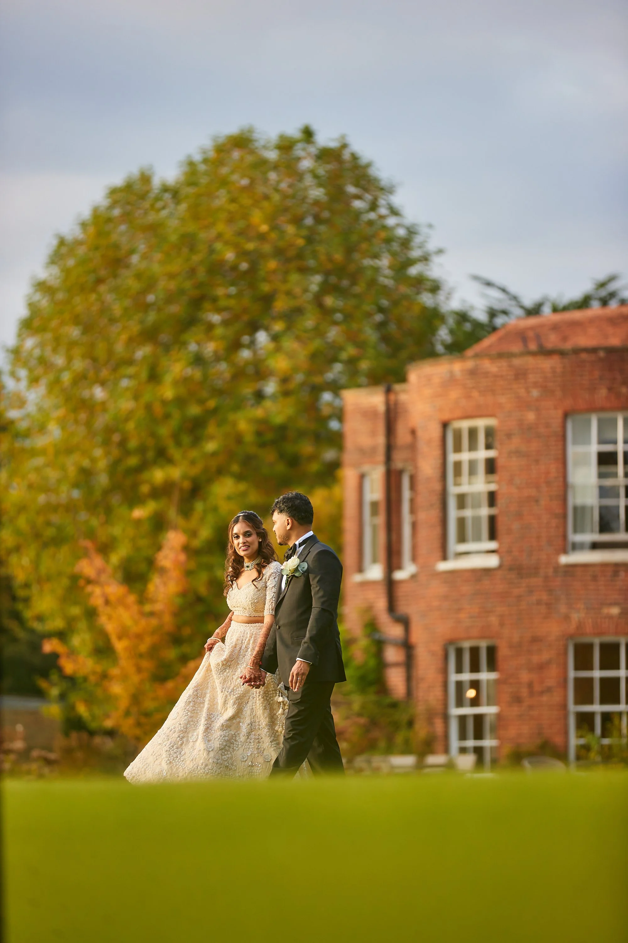 A bride and groom walking outdoors on their wedding day, with a brick building and autumn trees in the background.