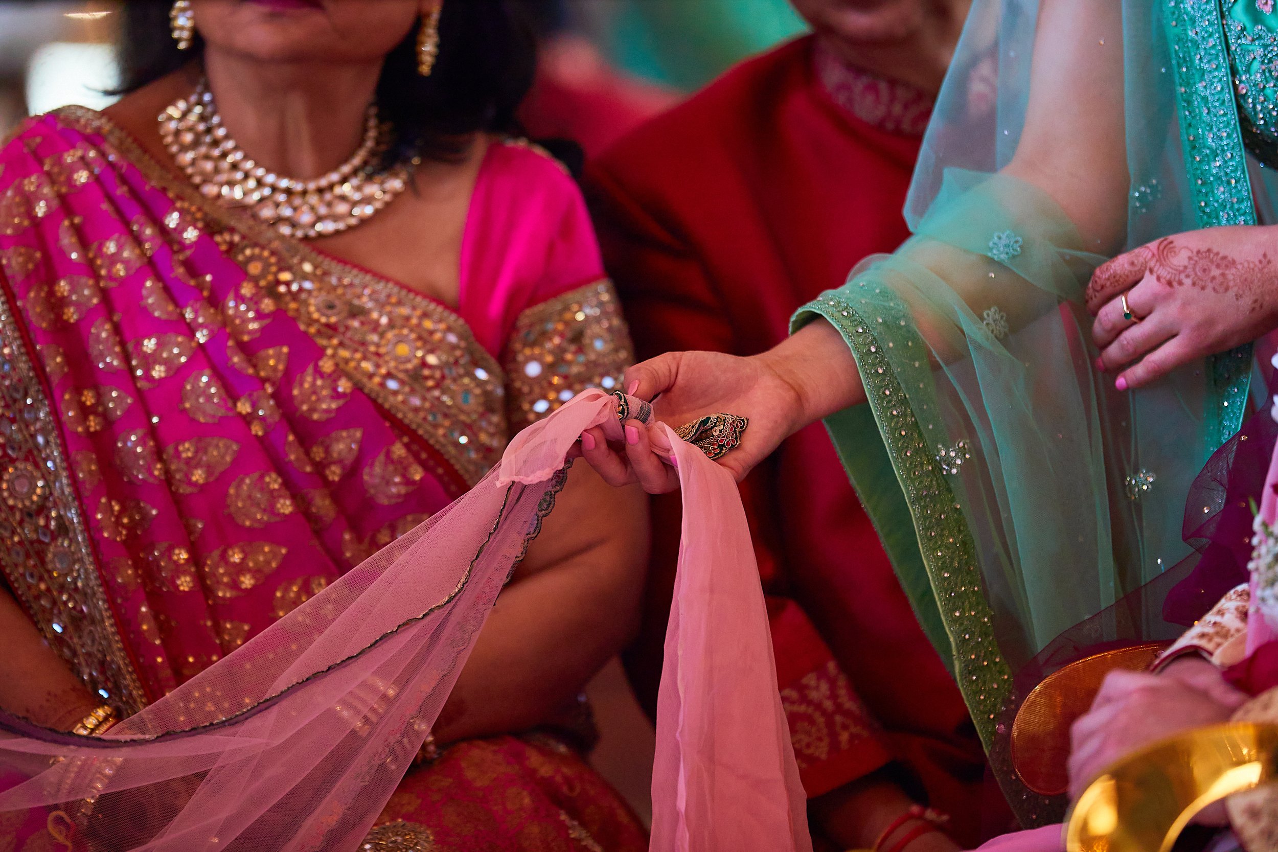 Close-up of women in colorful traditional Indian attire during a ceremony, with one woman holding a pink cloth and another touching her arm.
