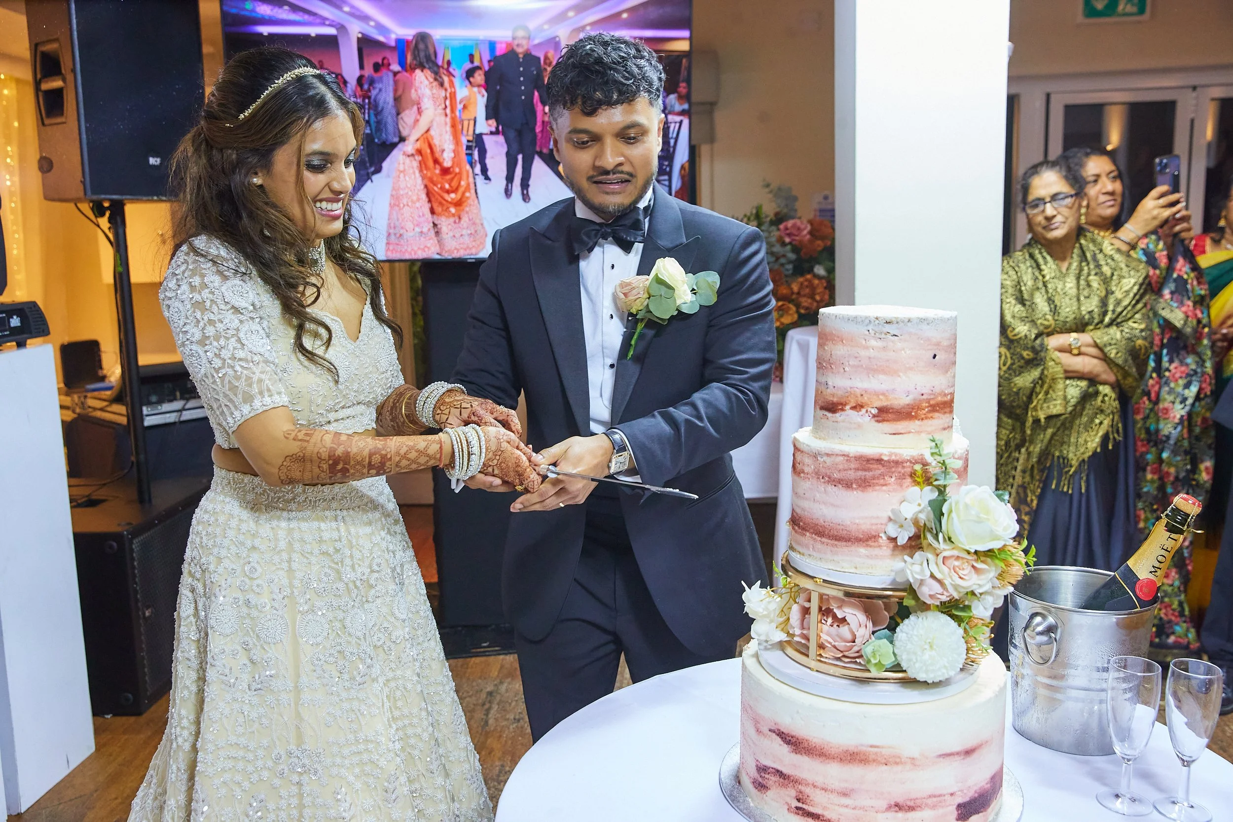 A newlywed couple cutting a wedding cake. The bride is wearing a cream-colored, embroidered dress with henna on her hands, and the groom is in a black tuxedo. The wedding cake is three-tiered with pinkish and white marbled frosting and floral decorat