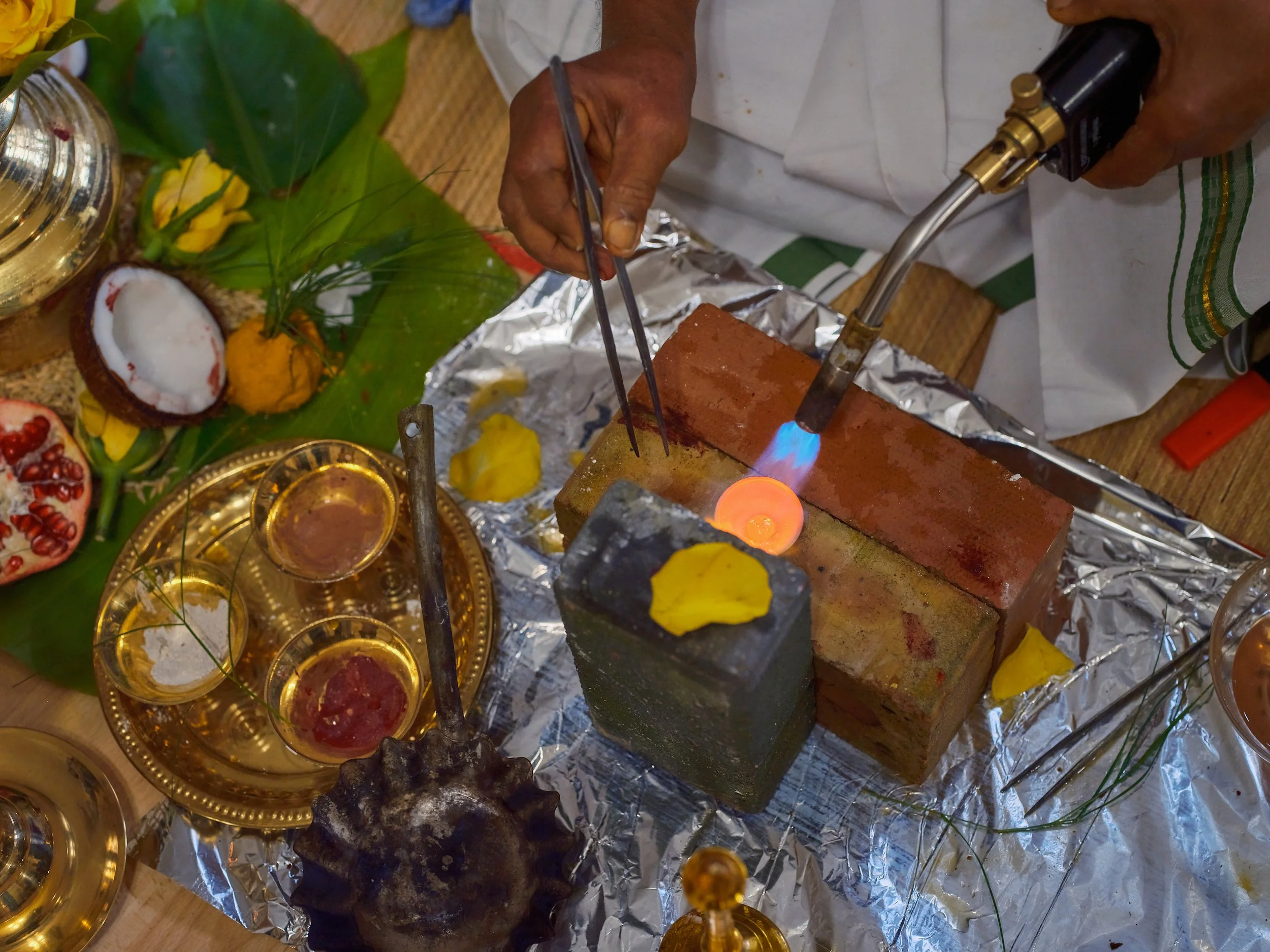 Person performing a ritual with clay lamps and candles on a silver-foil-covered surface, surrounded by flowers and offerings.