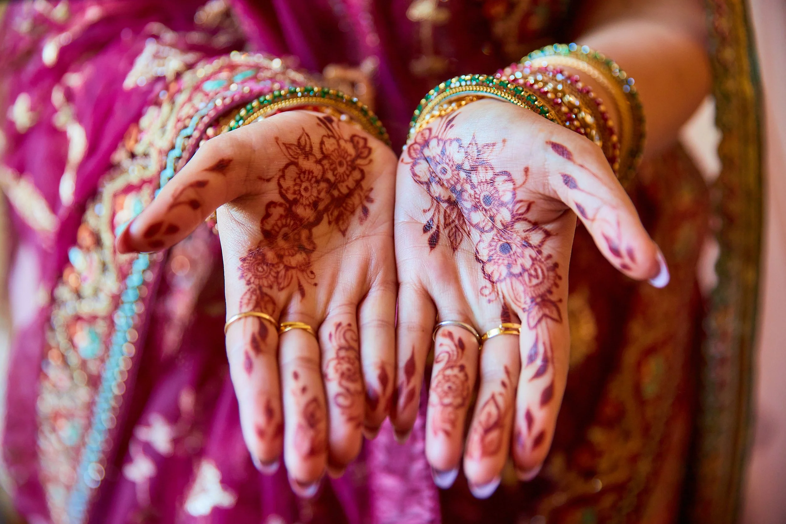 Close-up of a woman's hands with intricate henna designs, wearing gold and silver rings and colorful bangles, and dressed in traditional attire with detailed embroidery.