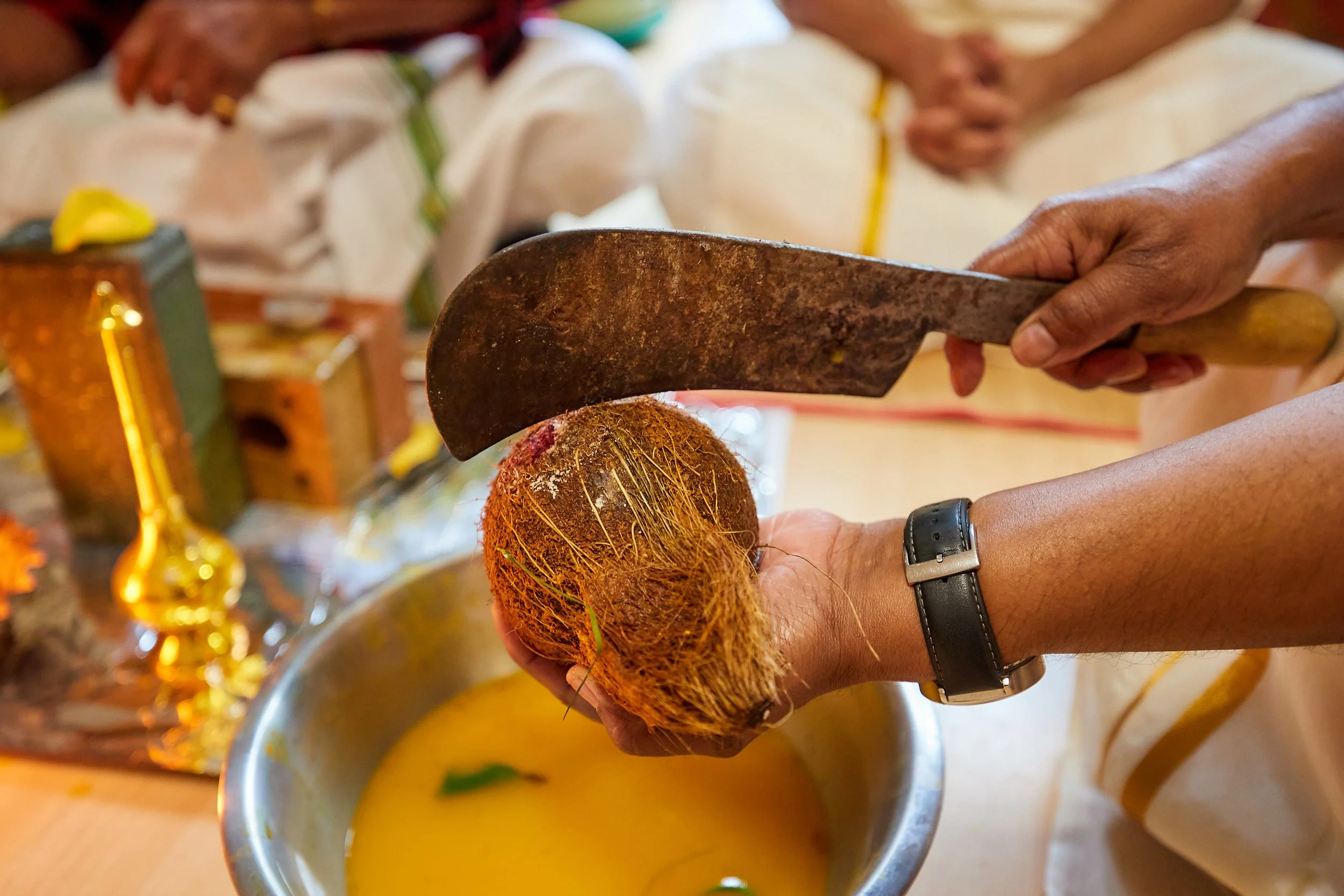 Person holding a coconut with one hand and holding a large kitchen knife with the other hand, preparing to open the coconut over a silver bowl filled with yellow liquid.