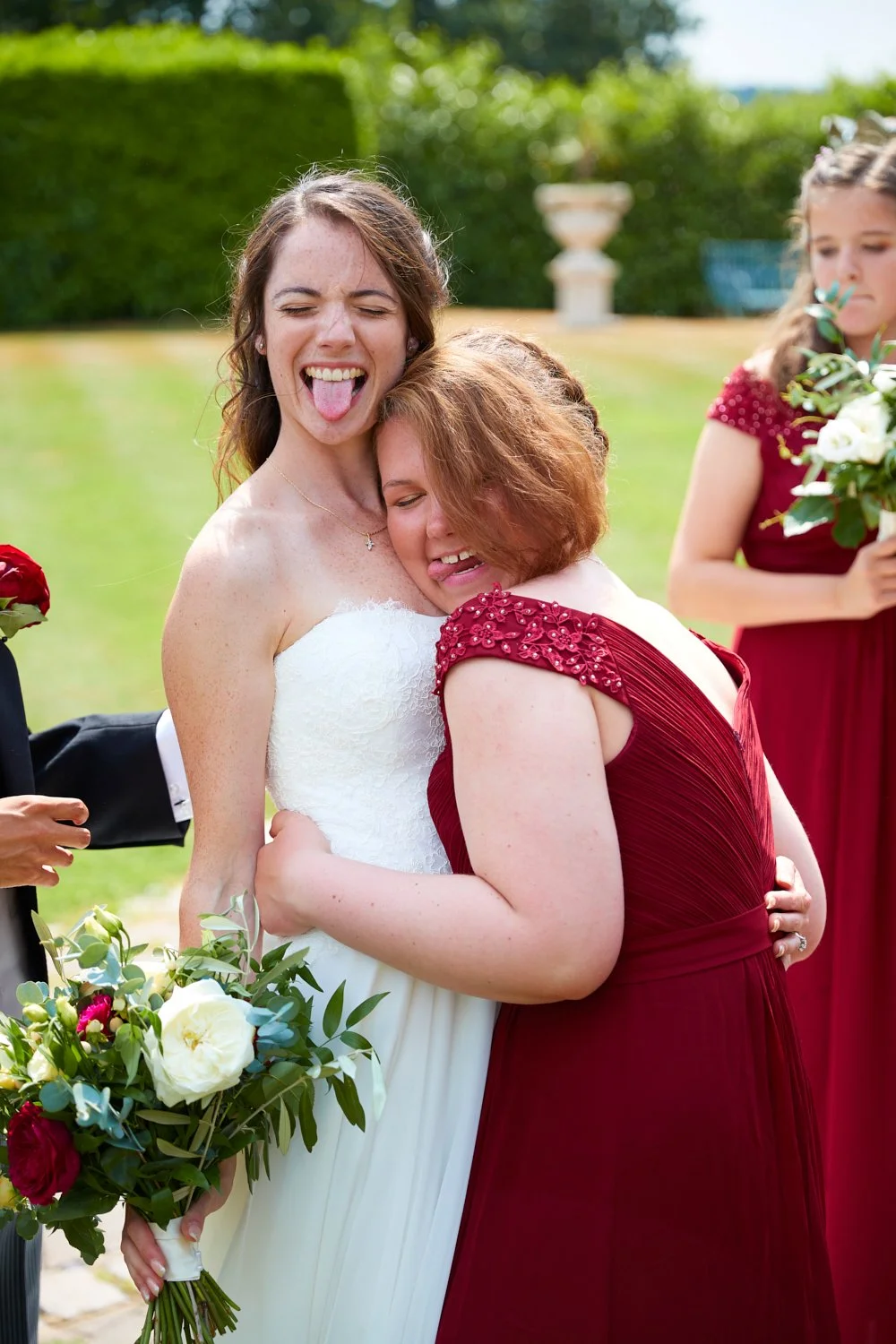 Two women, one in a white wedding dress and the other in a red dress, hugging and smiling at a wedding ceremony outdoors, with the bride sticking her tongue out playfully.