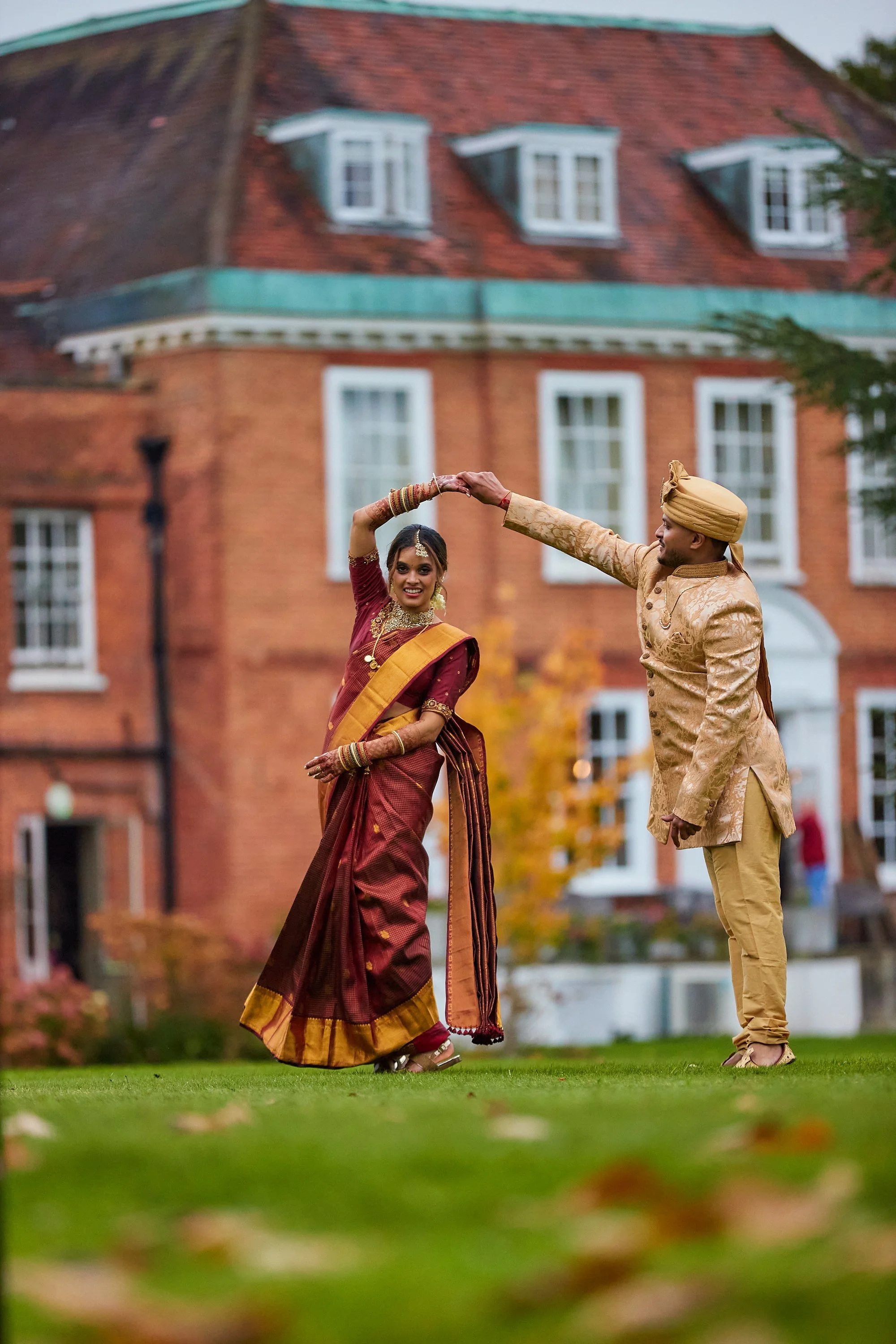 Indian couple dancing outdoors in traditional attire, with a large brick mansion in the background.