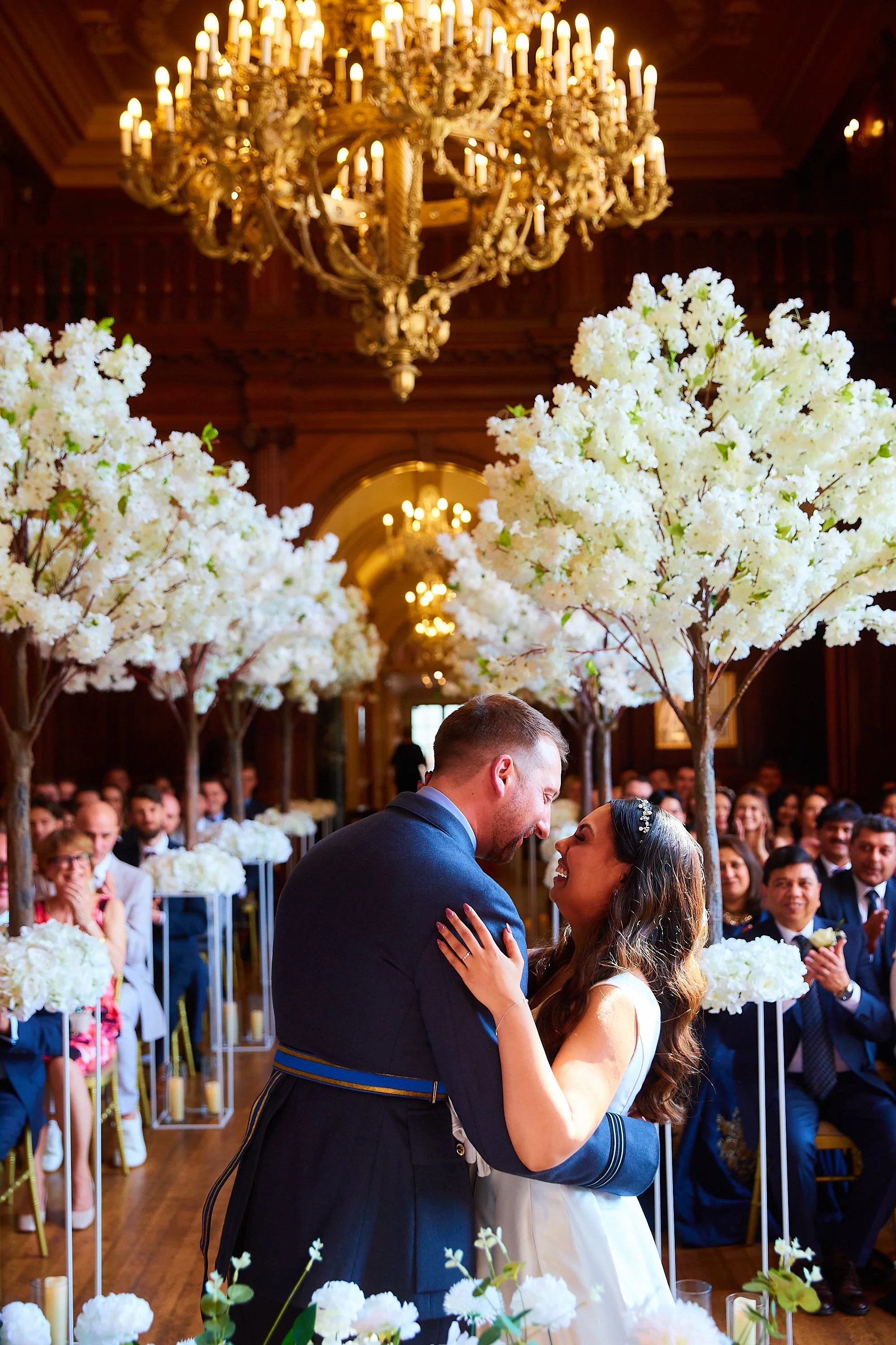 A wedding ceremony with a groom and bride looking at each other and smiling, surrounded by white flowers and guests clapping indoors with an ornate chandelier overhead.