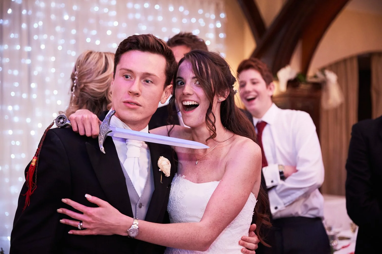 Scene from a wedding reception with a man holding a sword to his neck, bride smiling nearby, and guests laughing in the background.