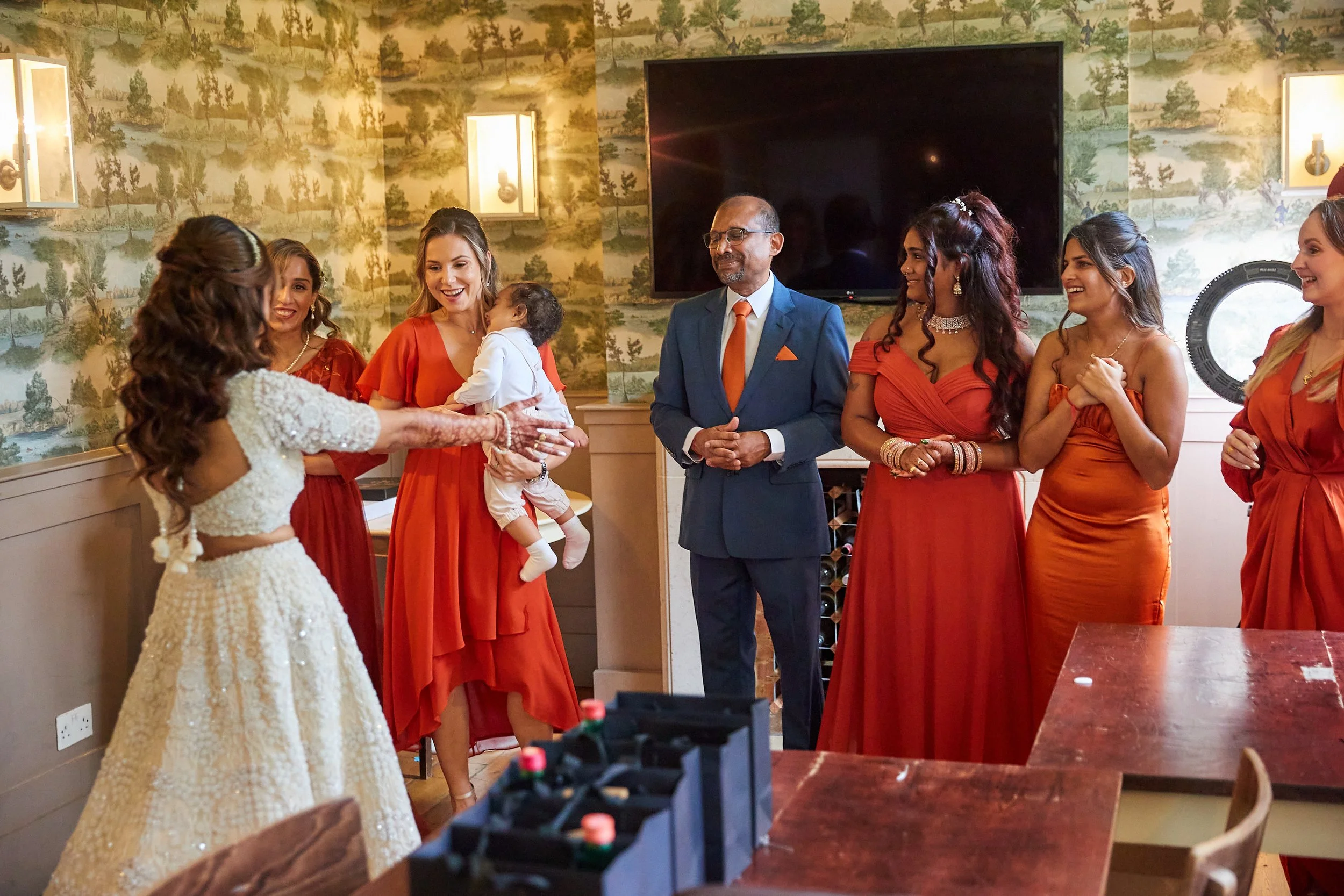 A group of women and a man are standing in a room, watching a woman in a white dress hold a baby. The women are dressed in red or orange dresses, and the man is in a blue suit with an orange tie. The setting appears to be a celebration or special occ