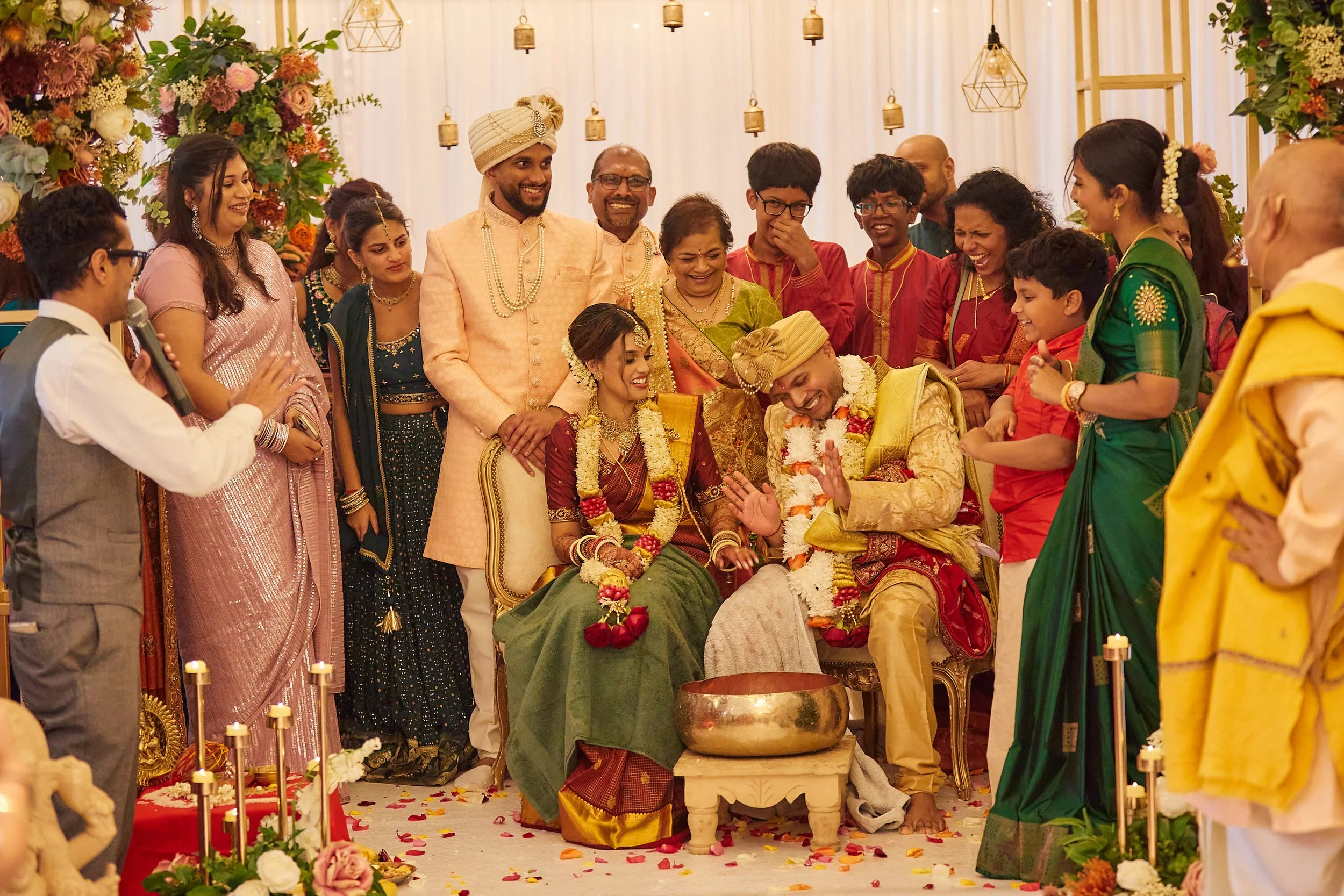Indian wedding ceremony with the bride and groom seated, wearing traditional attire and floral garlands, surrounded by family and friends celebrating and smiling.