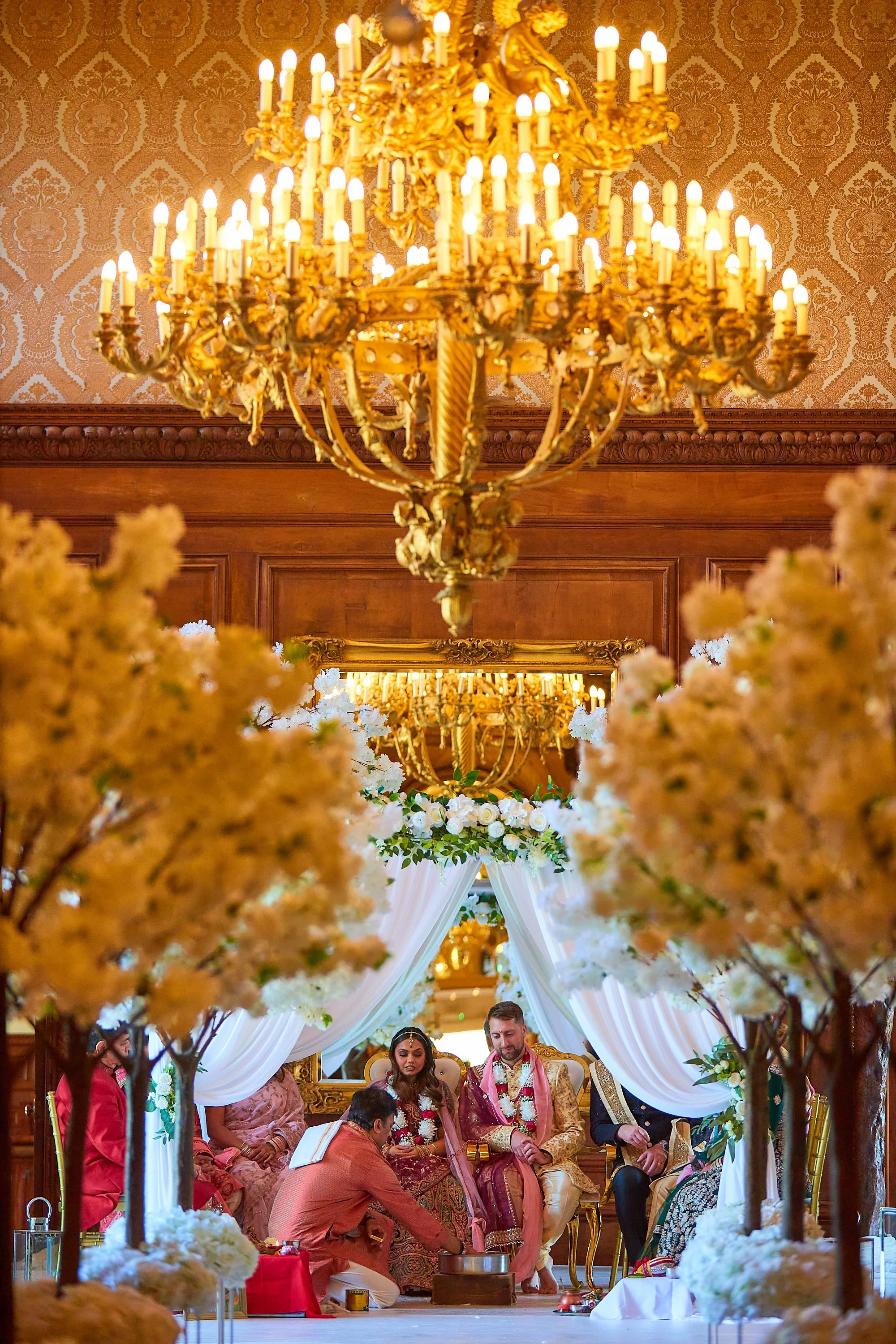 A wedding ceremony with a bride and groom seated on a decorated stage, surrounded by floral arrangements and illuminated by ornate chandeliers.