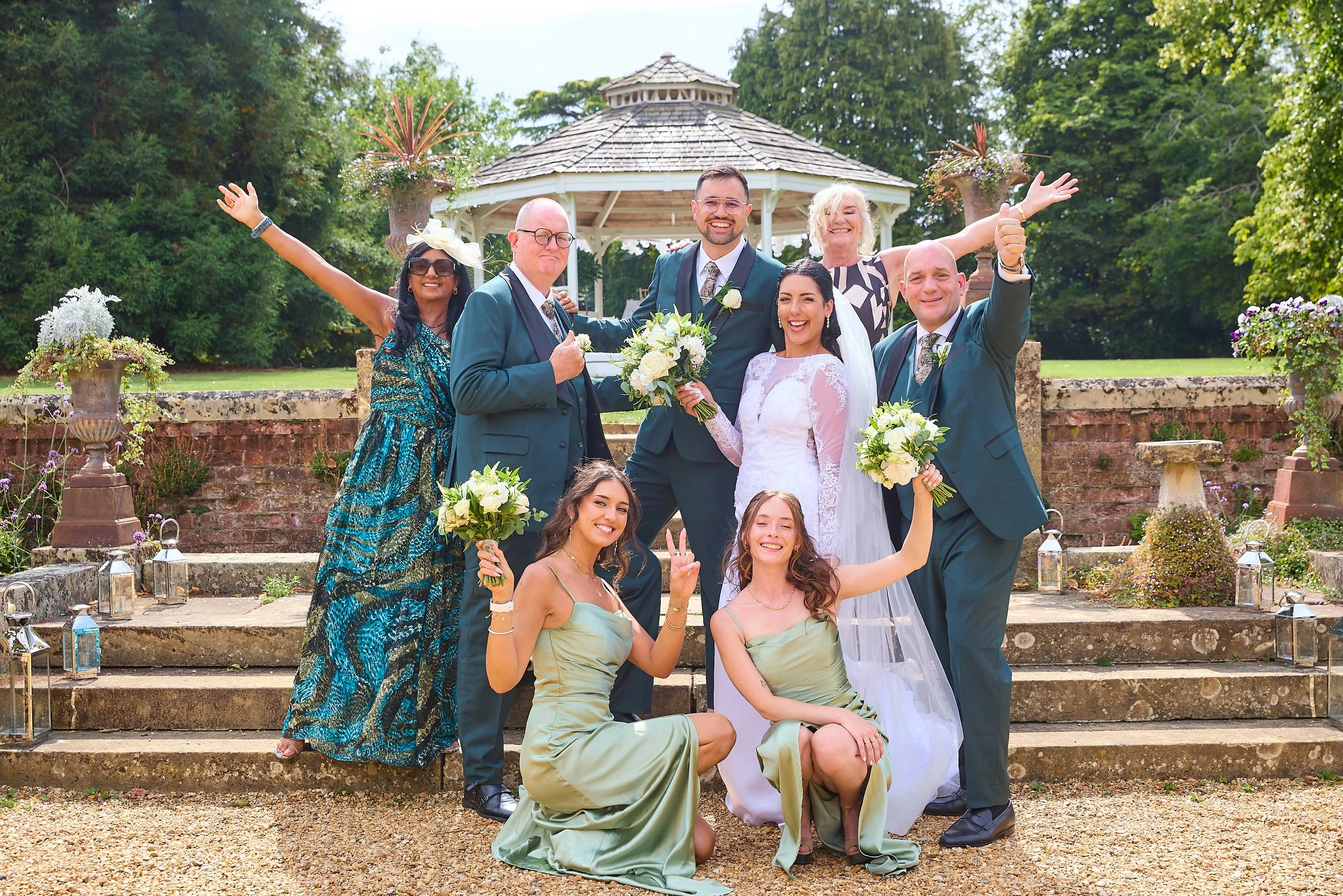 A group of people dressed in wedding attire celebrating outdoors on steps with a gazebo in the background. The bride and groom are in the center, surrounded by family and friends holding bouquets of white flowers.