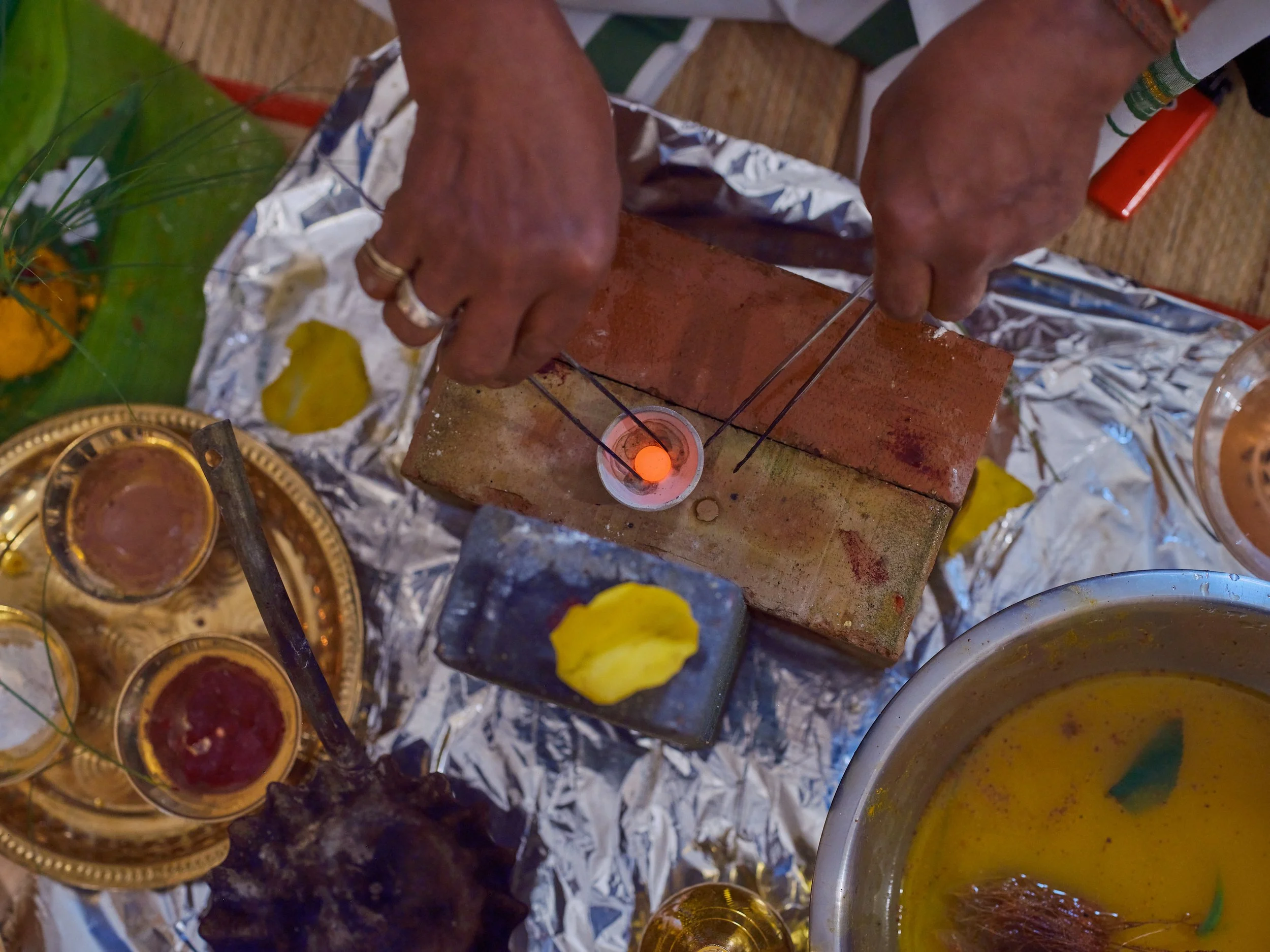 A person performing a religious ritual involving a small fire on a brick with two metal sticks, surrounded by offerings and yellow flowers.
