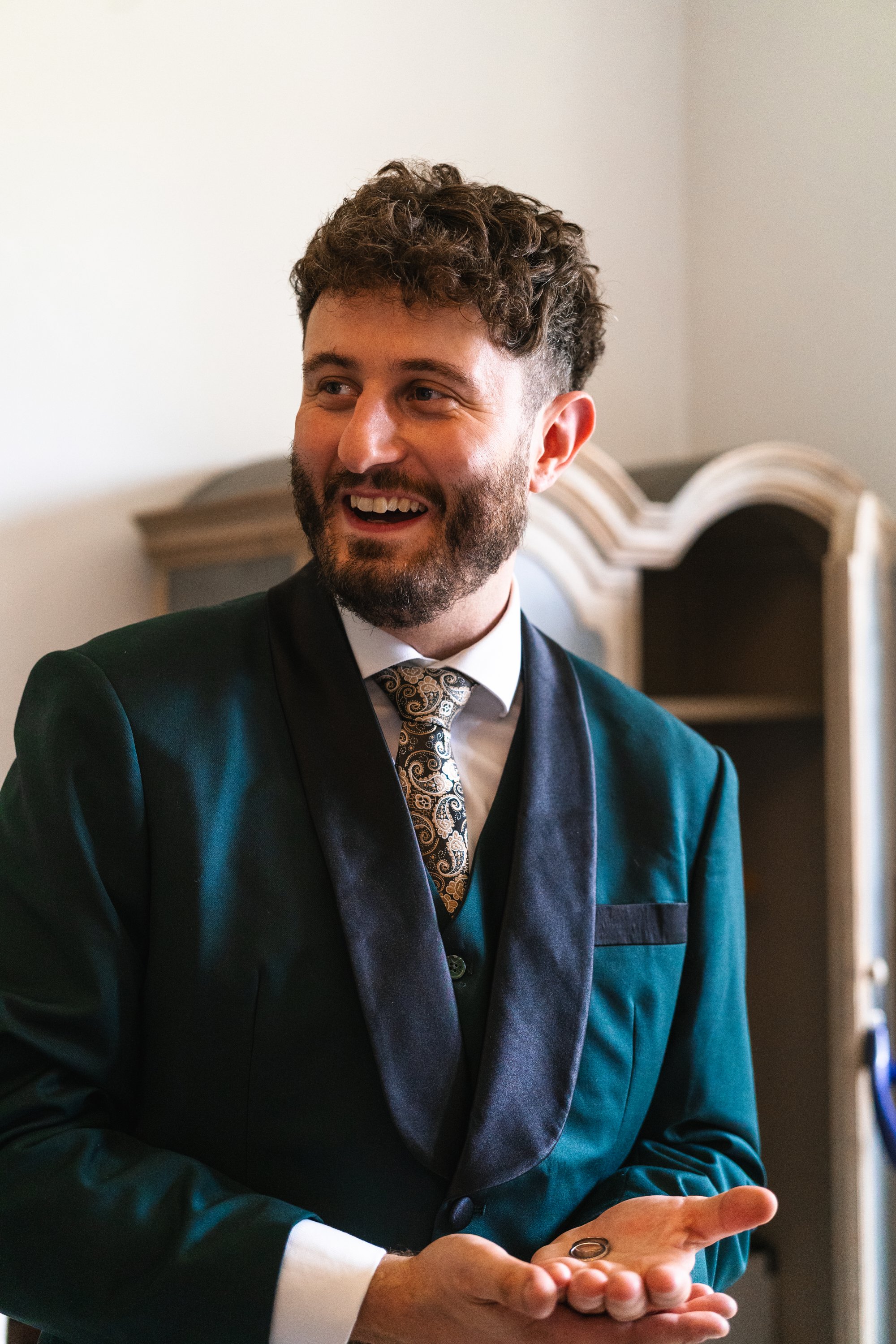 A man with a beard and curly hair dressed in a dark suit, white shirt, and patterned tie, smiling and holding wedding rings in his hand.
