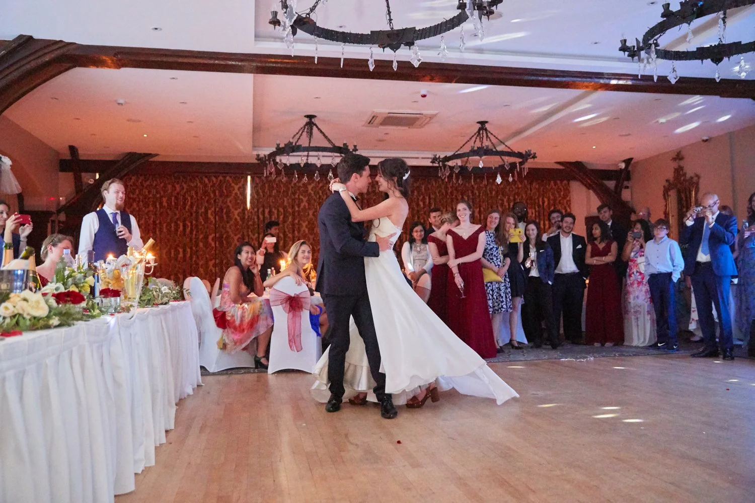 Couple dancing at wedding reception with guests watching and taking pictures in an elegant decorated hall.