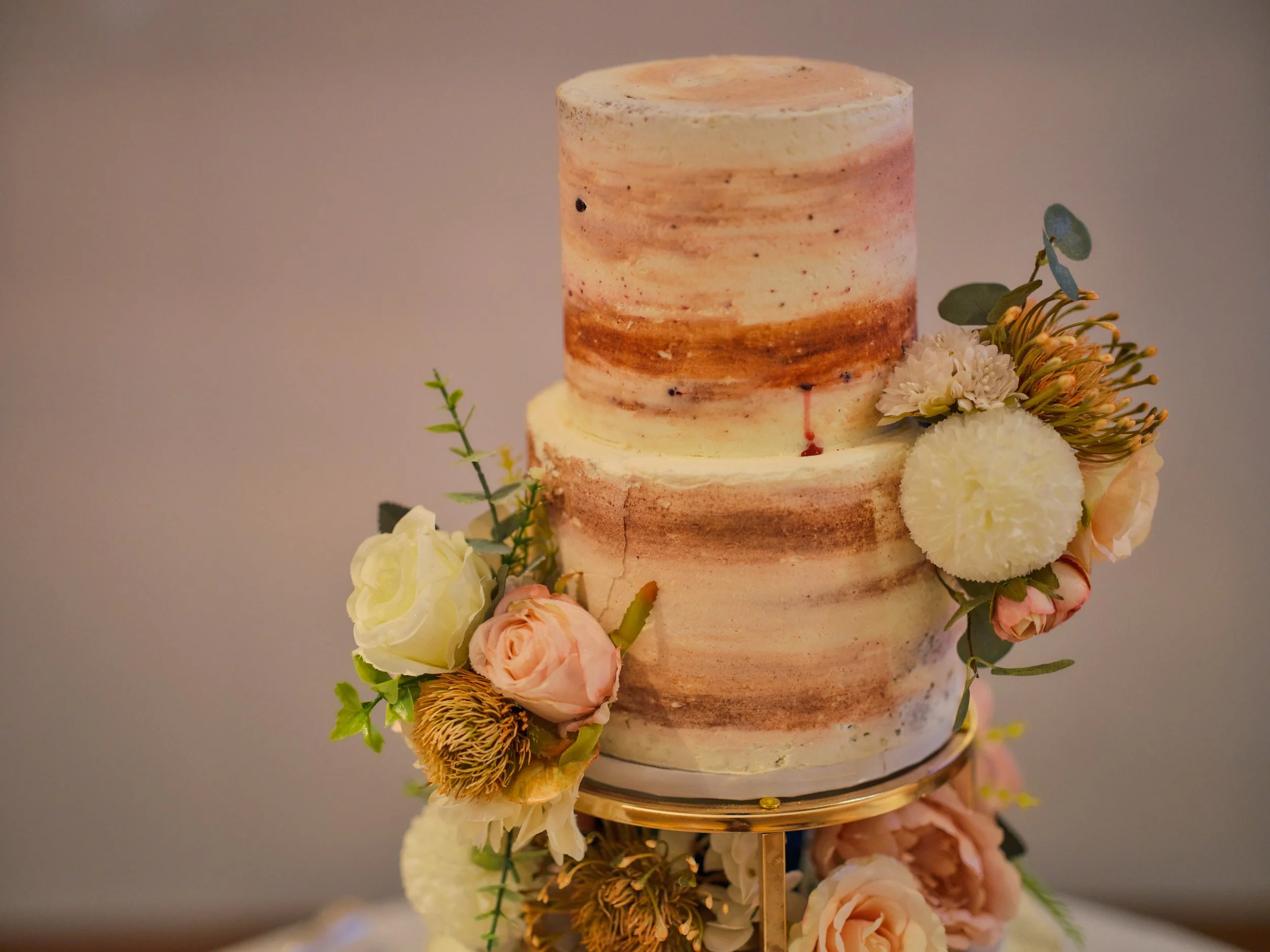 A two-tiered wedding cake with semi-naked buttercream frosting and a floral arrangement featuring roses and greenery.