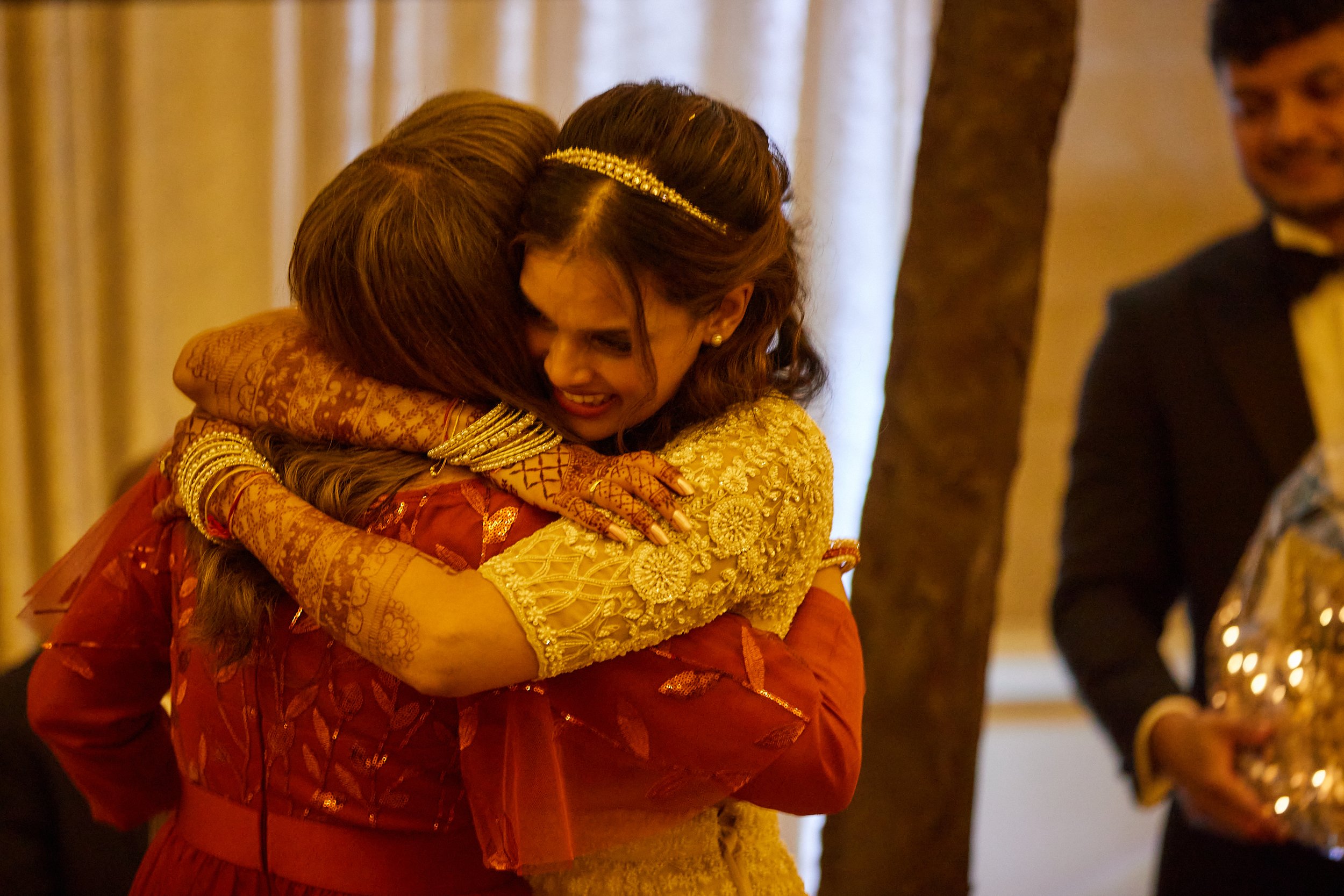 Two women hugging at a celebration. One woman wears a yellow lace dress and a tiara, while the other woman is dressed in red with intricate henna on her hands. A man in a tuxedo is in the background holding a tray.