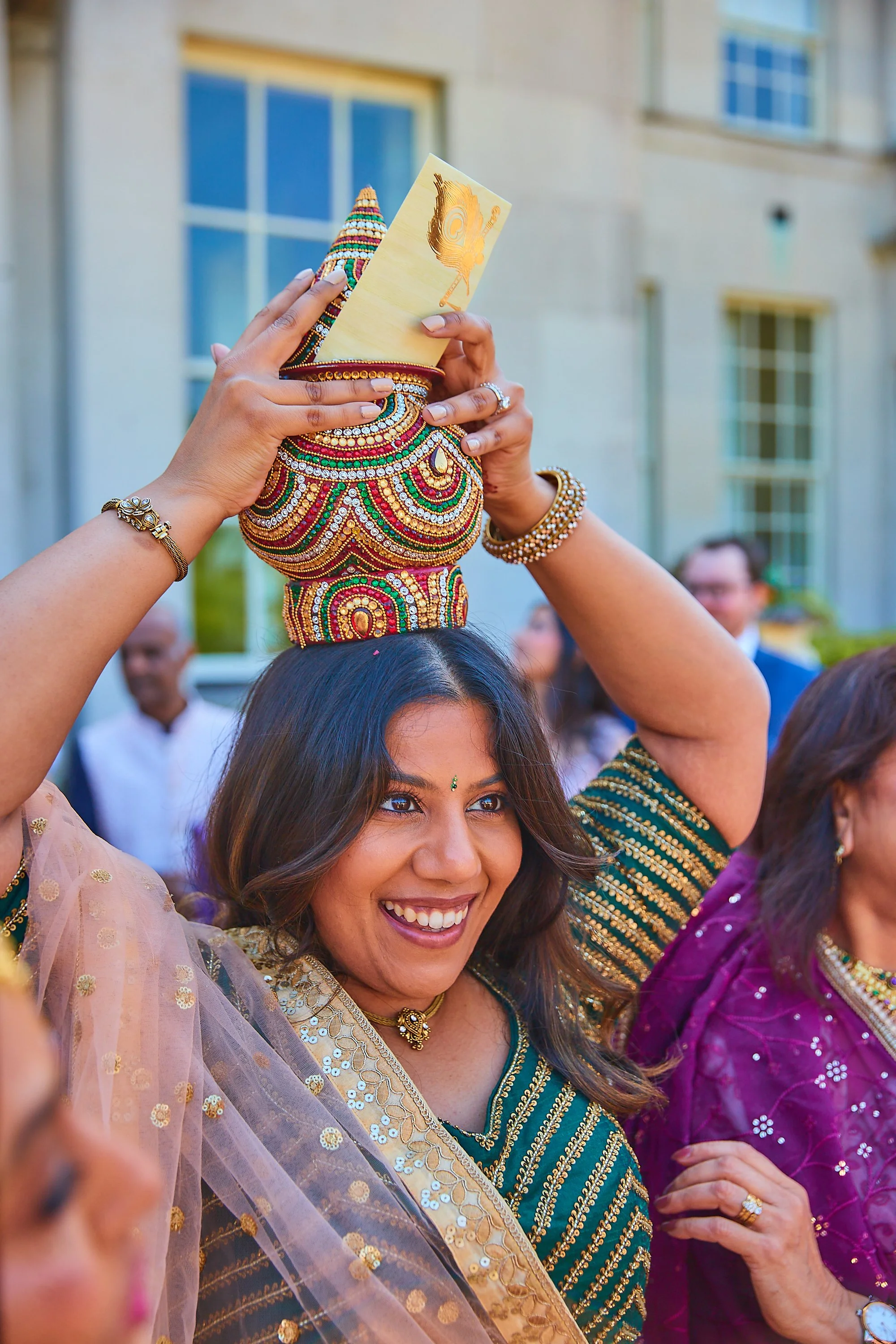 A woman smiling and wearing a traditional dress, balancing a colorful, beaded pot on her head during a celebration, with other people in festive attire in the background.