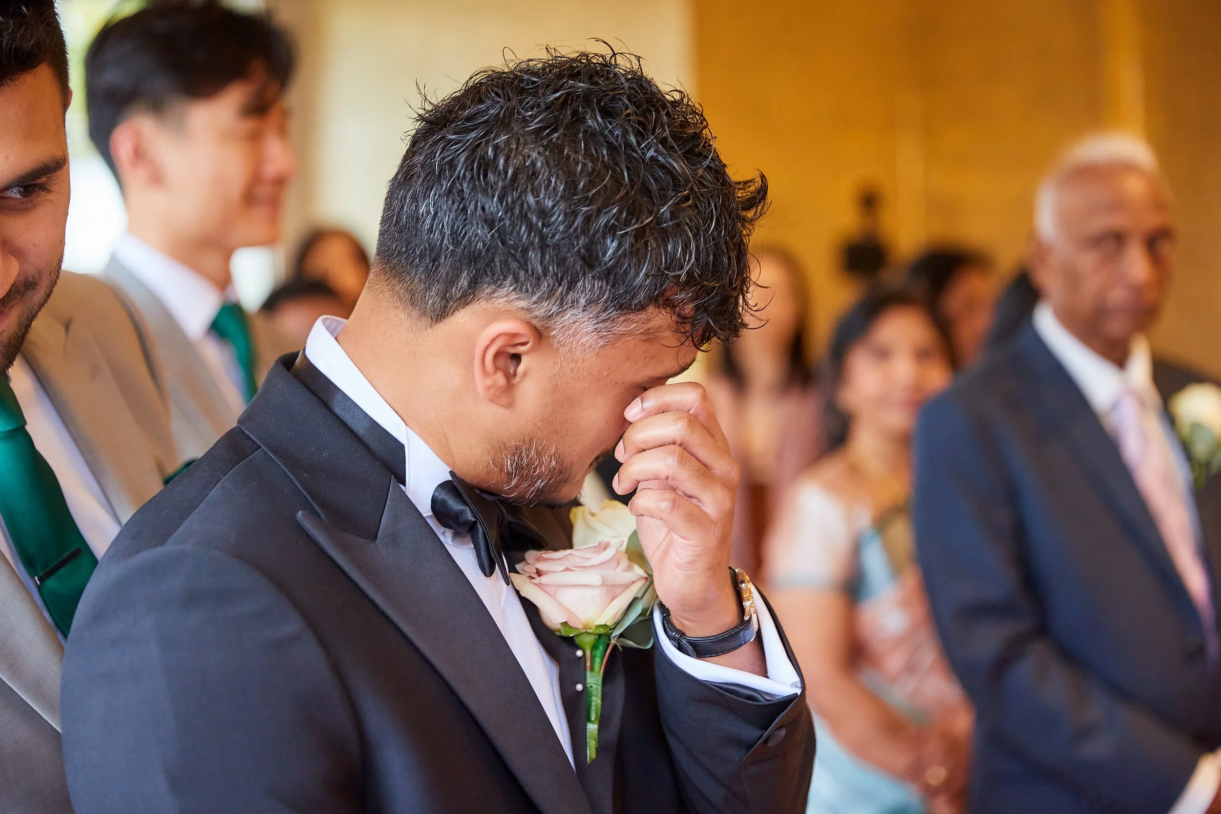 Man in tuxedo with a pink rose boutonniere, tears in eyes, holding his face in his hand during a wedding ceremony.