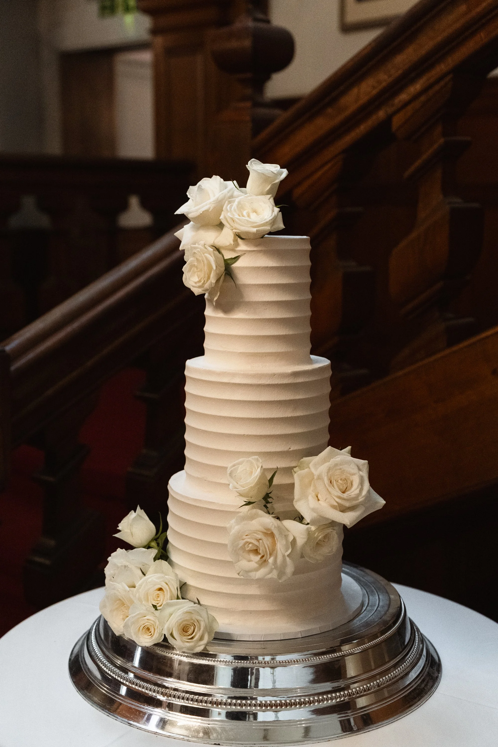 A tall, white wedding cake with horizontal ribbing, decorated with white roses, placed on a silver cake stand.