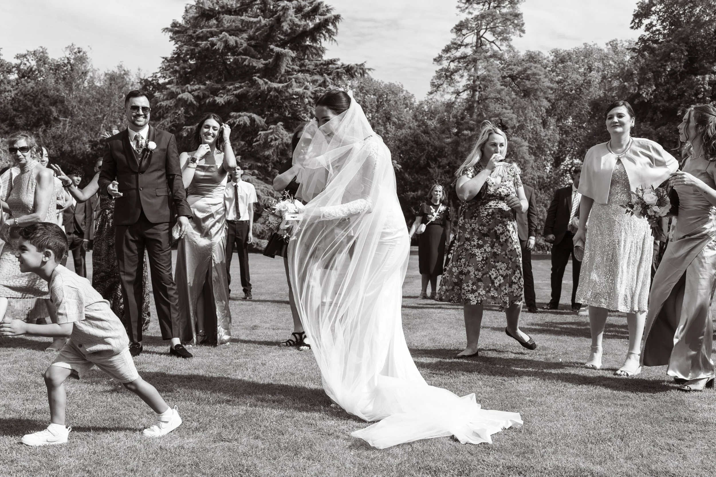 A group of people gathered outdoors, including a bride in a wedding dress and veil holding a bouquet, surrounded by guests in formal attire, on a grassy area with trees in the background.