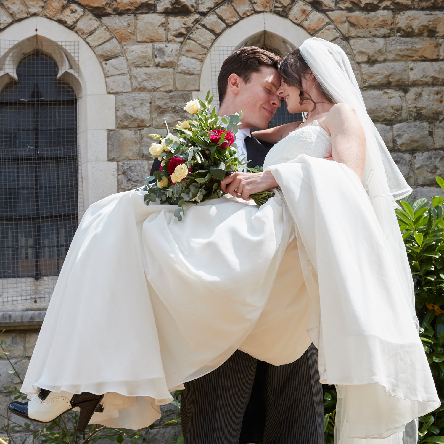 A bride and groom sharing a romantic moment outside a stone church. The groom lifts the bride, who holds a bouquet of red and cream flowers, as they lean their foreheads together with eyes closed.