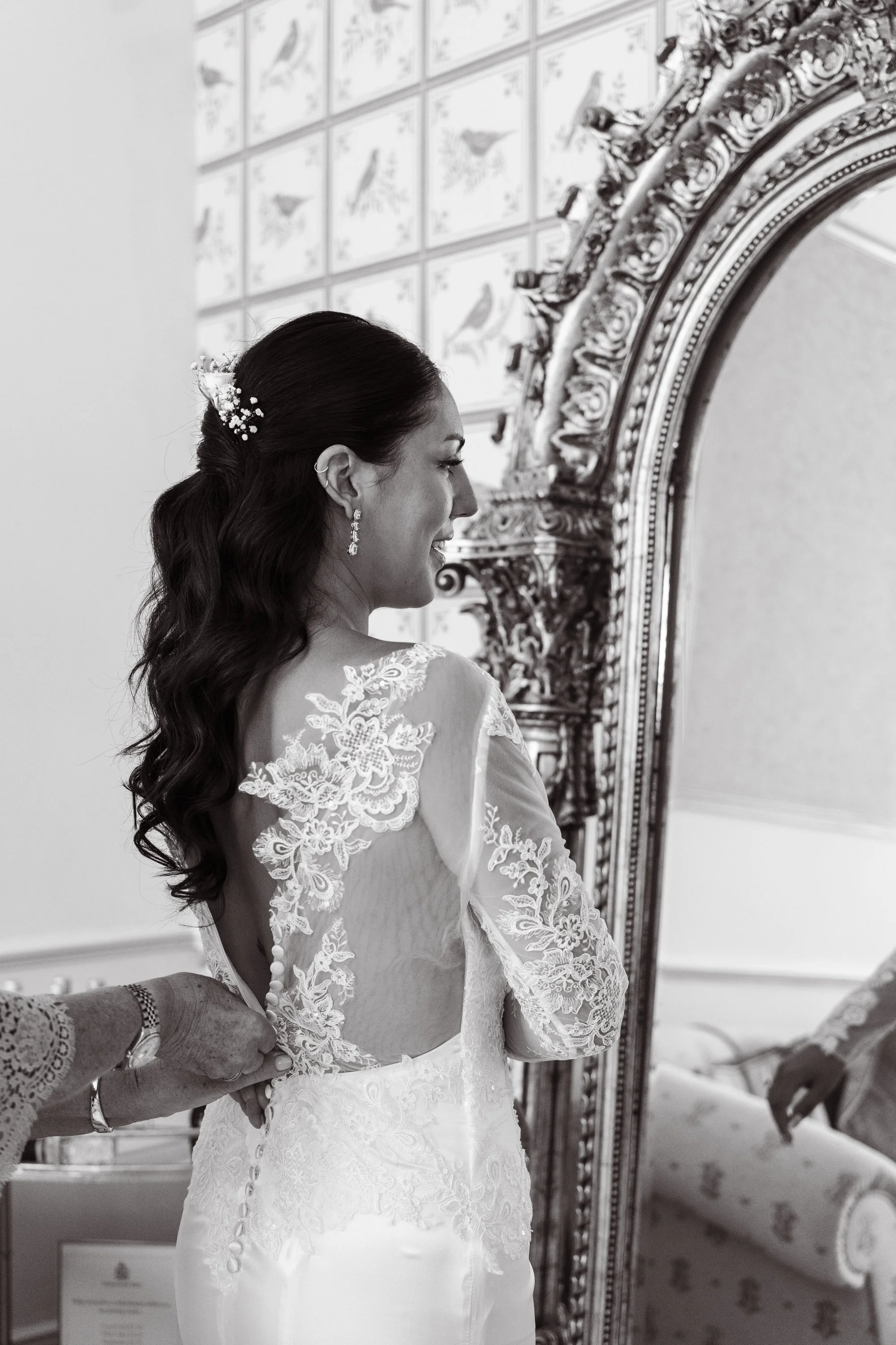 A bride is getting dressed in her wedding gown, with someone assisting her buttoned up the back. She has long dark hair styled in loose waves with a small floral hairpiece, and is wearing earrings. The scene is reflected in an ornate, large mirror.