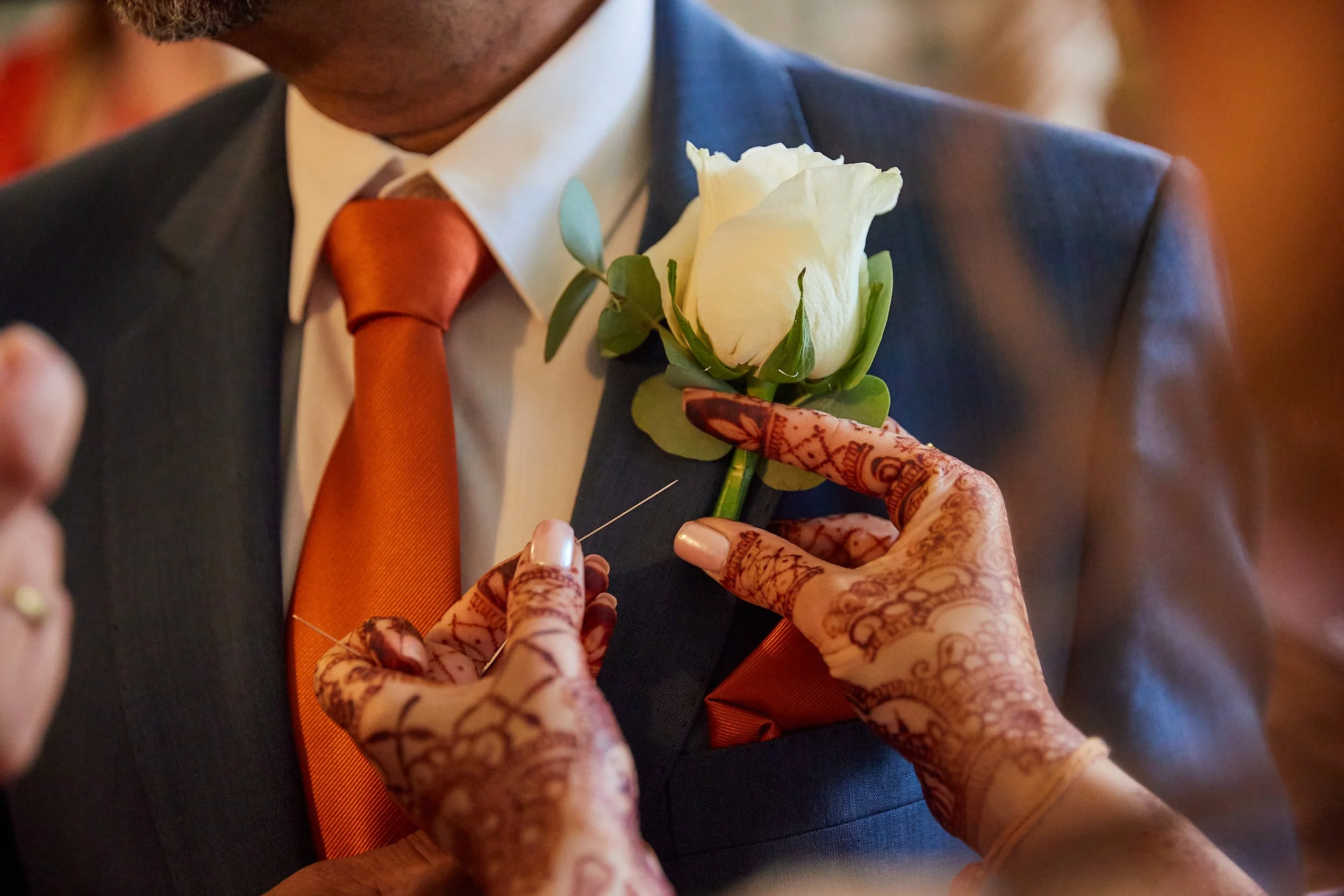 Close-up of a woman with intricate henna designs applying a white boutonniere to a man's suit lapel during a ceremony, possibly a wedding.