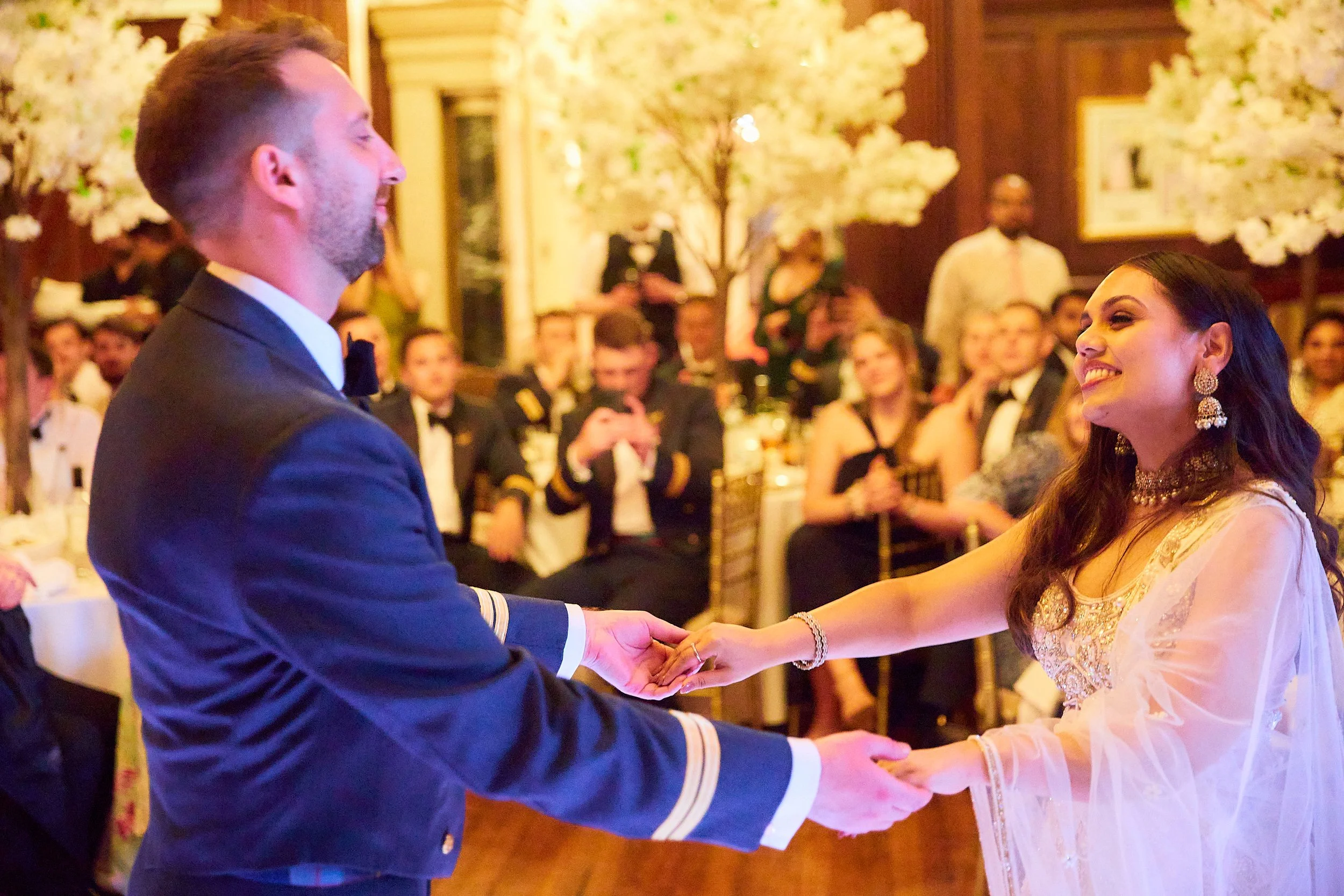 A couple dressed in wedding attire holding hands and smiling at each other during a wedding ceremony in a decorated reception hall with guests in the background.