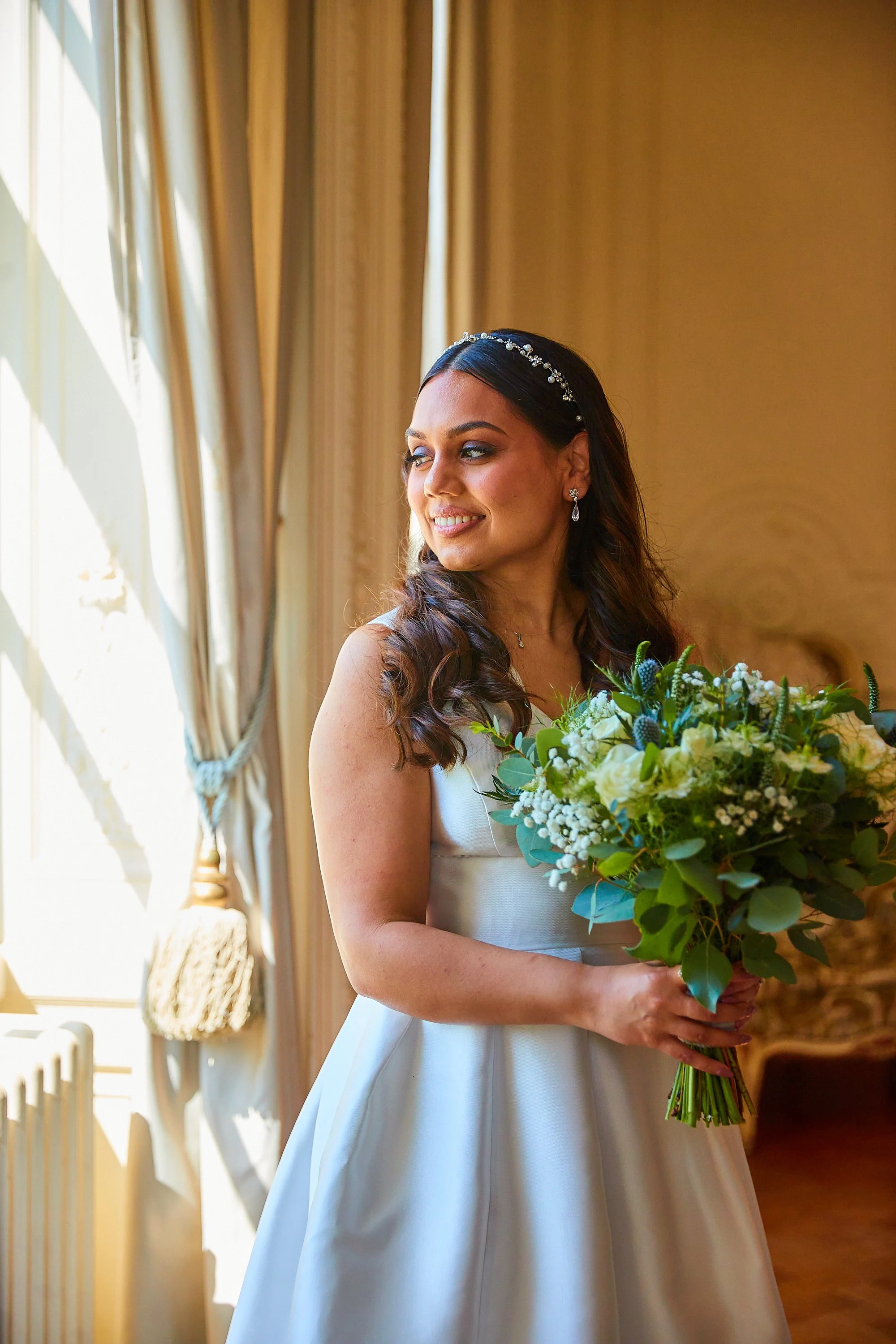 A bride standing indoors, holding a bouquet of white and green flowers, looking out a window with sunlight streaming in, in a wedding dress.