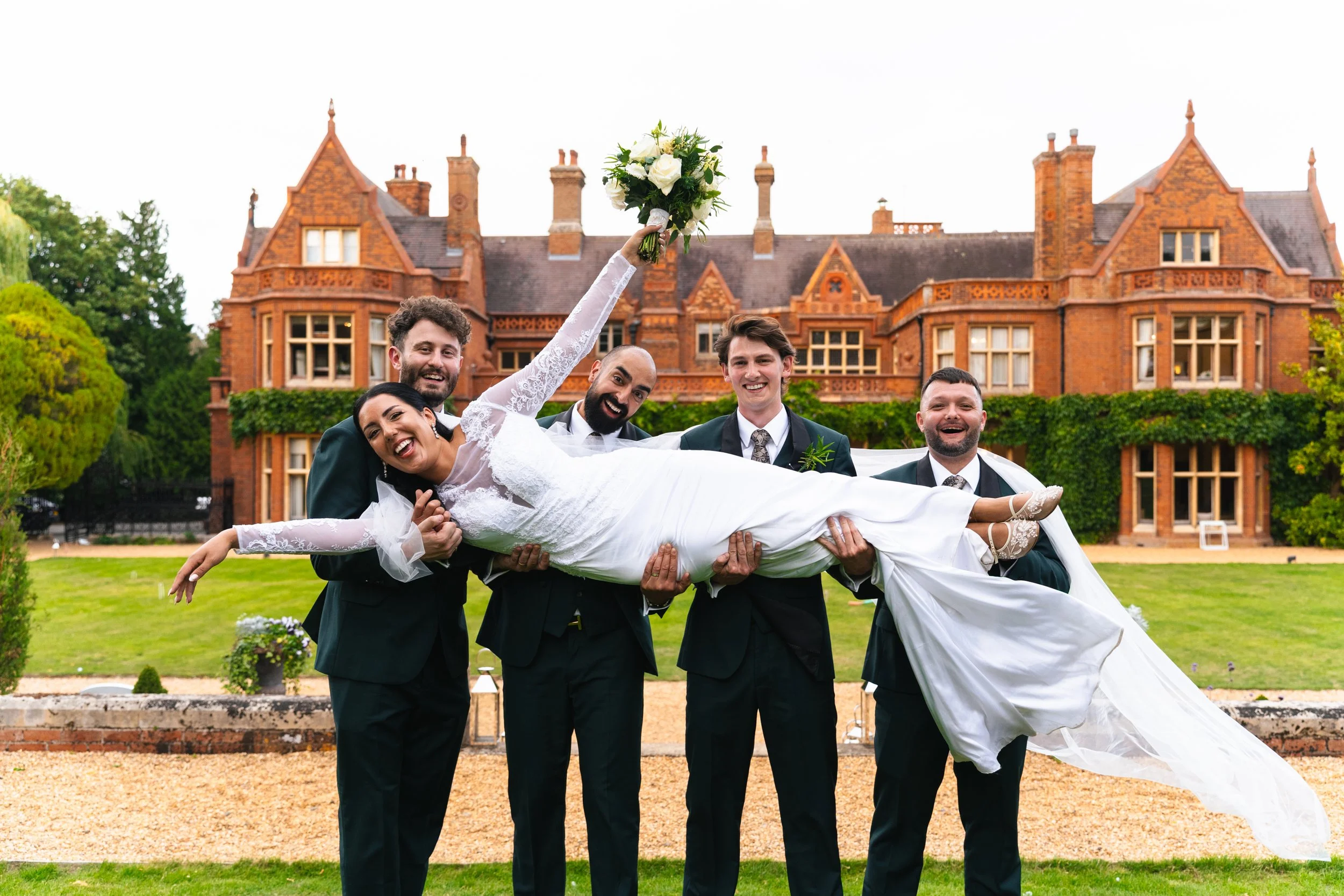 Bride in white wedding gown being lifted by four men in suits, holding a bouquet of flowers, in front of a large historic red brick mansion with green ivy and lawn.