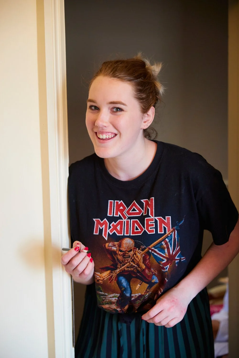A young woman with light skin and red hair smiling, wearing an Iron Maiden T-shirt, standing in a doorway, holding a pen.