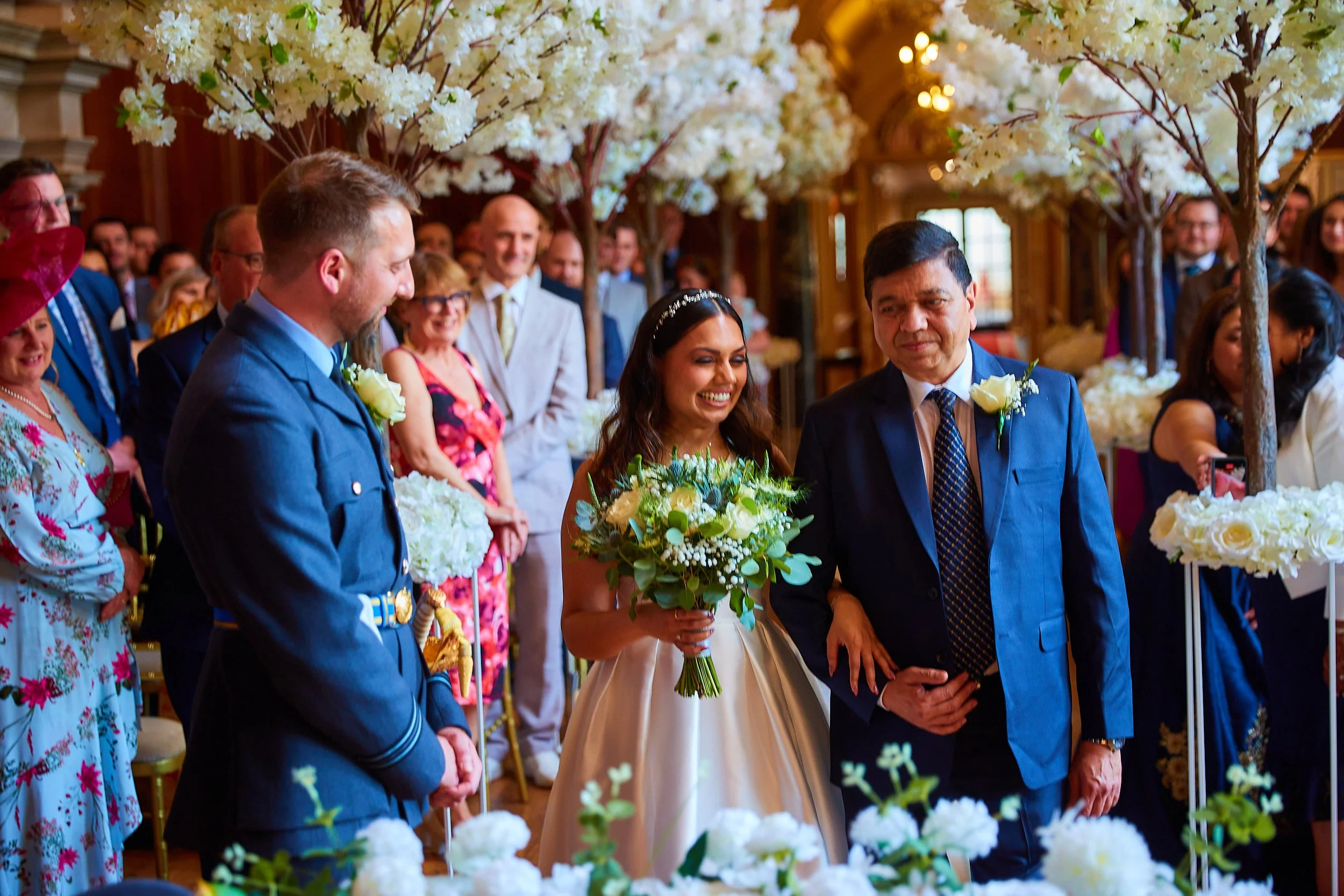 Bride in a white dress holding a bouquet being walked down the aisle at a wedding ceremony, surrounded by guests and floral decorations.